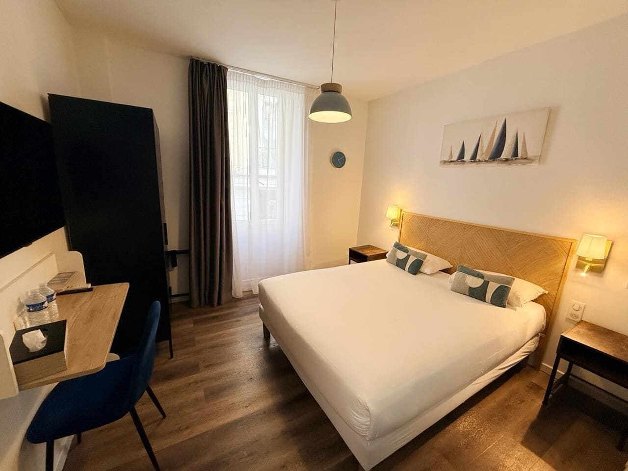 A hotel room with a bed, a desk, a chair, a nightstand, a lamp, and a window with curtains.