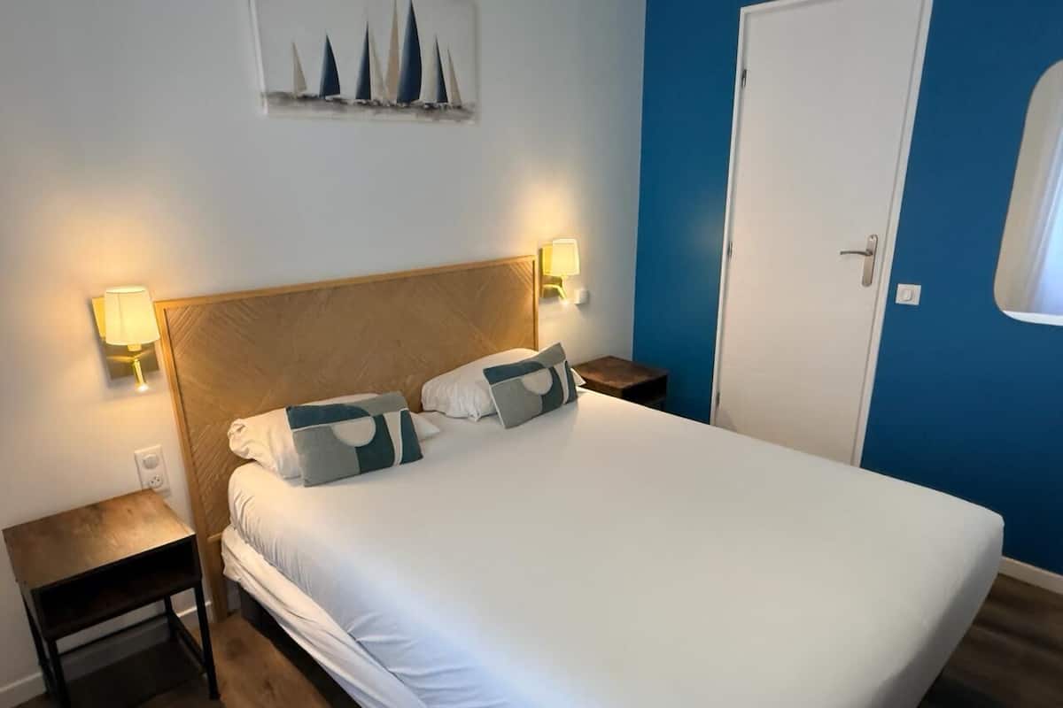 Ein ordentlich bezogenes Bett mit weißen Leinentüchern und zwei Kissen, begleitet von zwei Nachttischen mit Lampen, in einem Zimmer mit blauen Wänden und einem hölzernen Kopfteil.