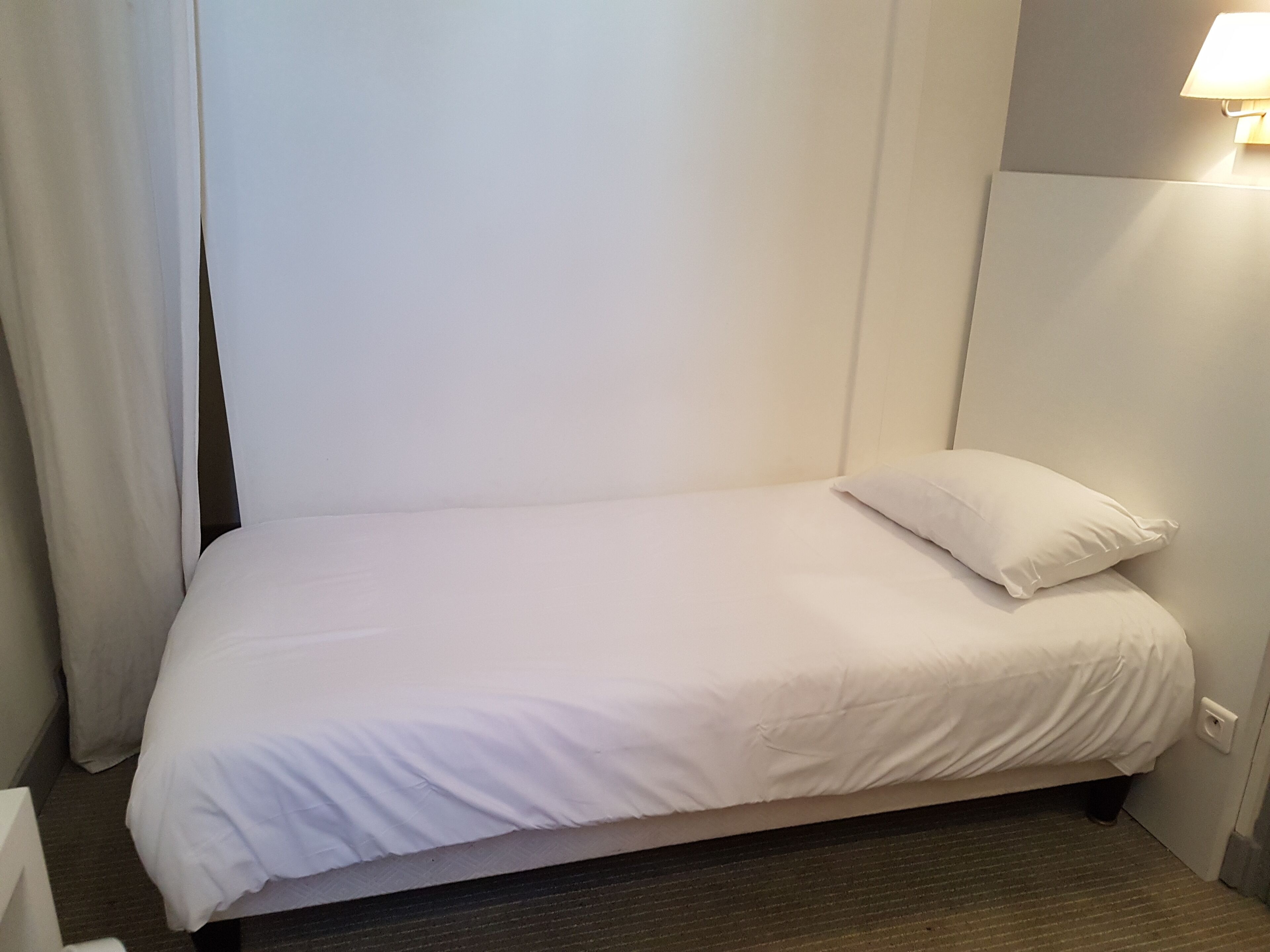 A single bed with white bedding in a room with a plain white wall and a light fixture above it.