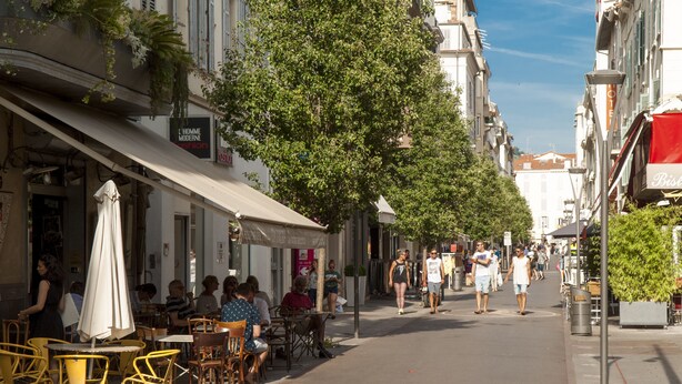 Une rue avec des terrasses, un arbre et des bâtiments portant des enseignes.