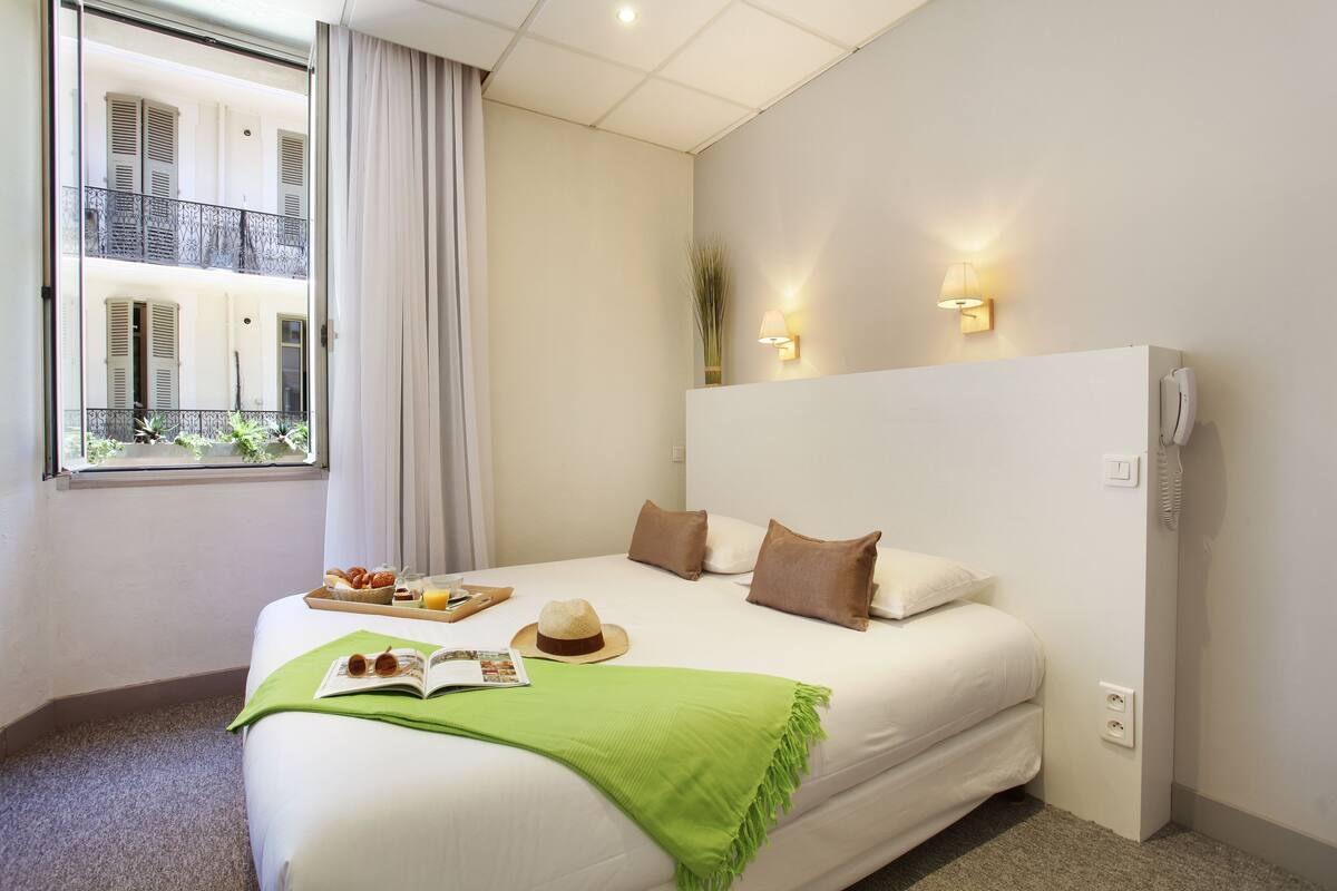 Ein Hotelzimmer mit Bett, Fenster mit Vorhängen, Balkonblick und Nachttisch mit Tablett mit Essen und Hut.