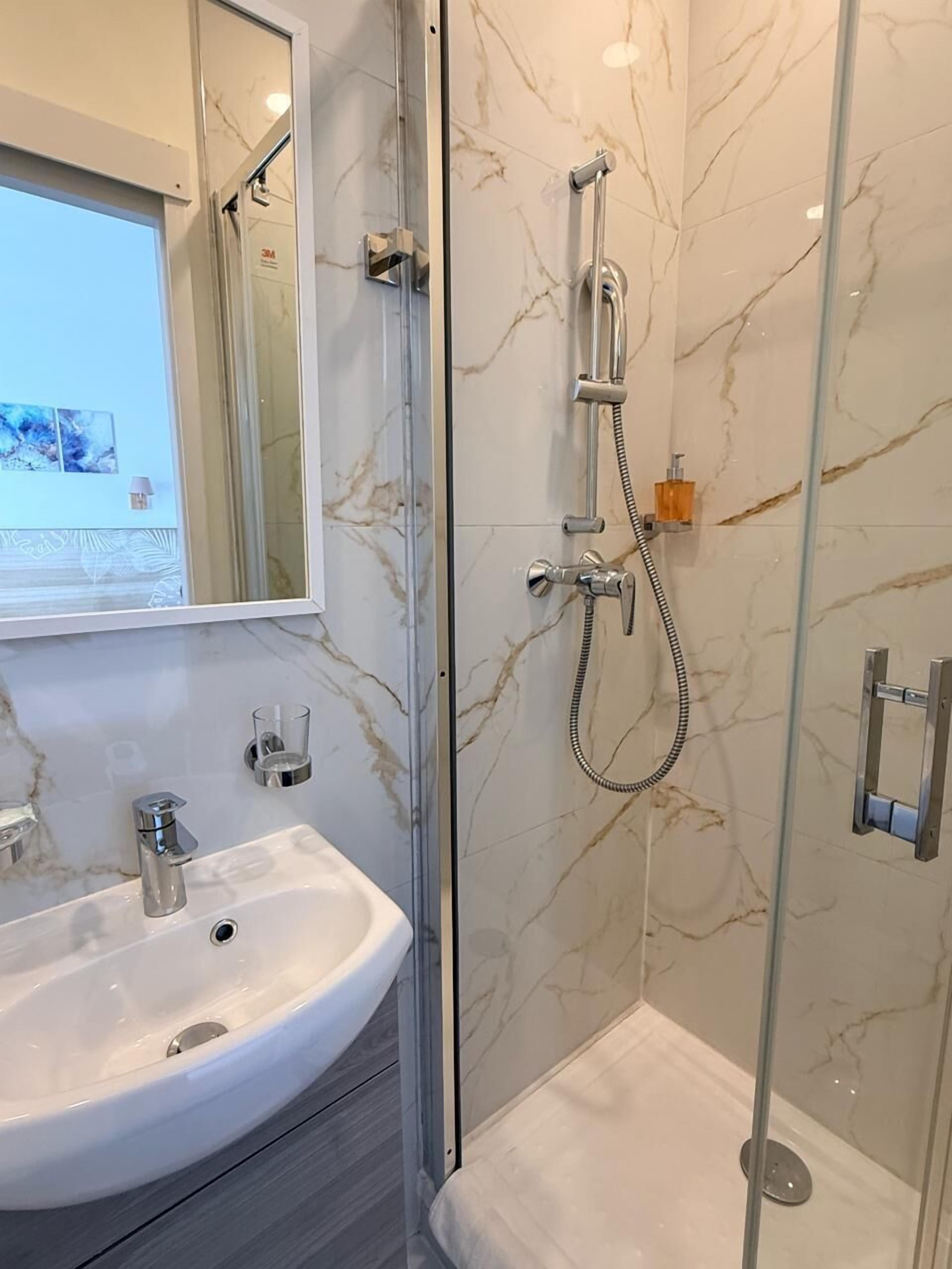 A bathroom with a marble wall, a glass shower enclosure, a white sink, and a mirror.