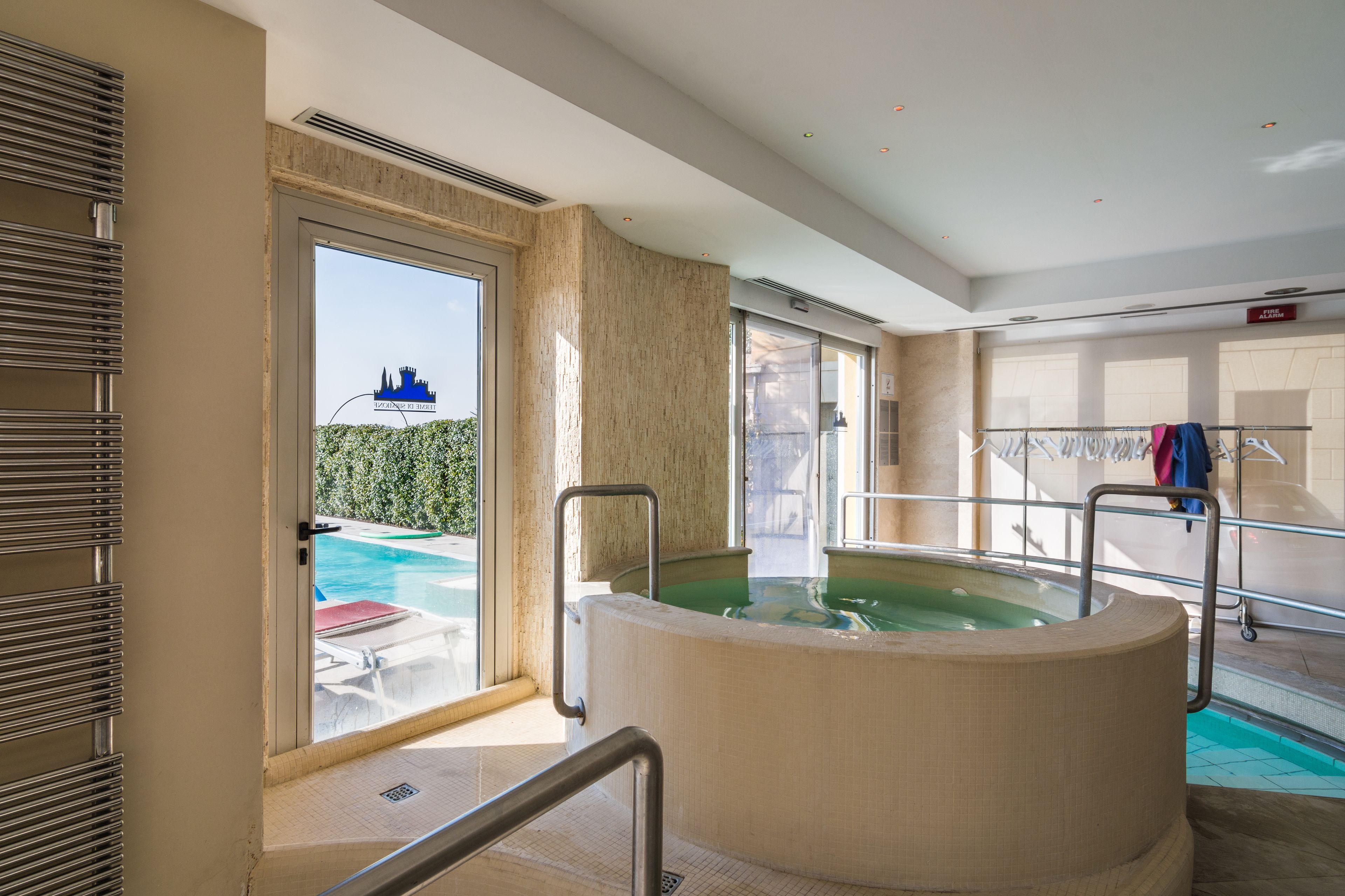 A modern indoor pool area with a large hot tub, a glass door leading outside, and a clothing rack with towels.