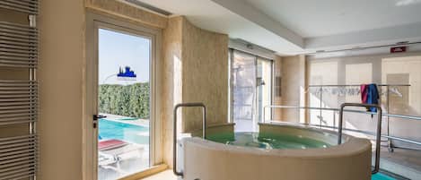 A modern indoor pool area with a large hot tub, a glass door leading outside, and a clothing rack with towels.