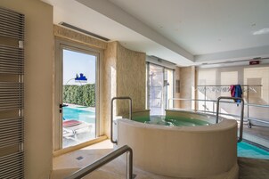 A modern indoor pool area with a large hot tub, a glass door leading outside, and a clothing rack with towels.