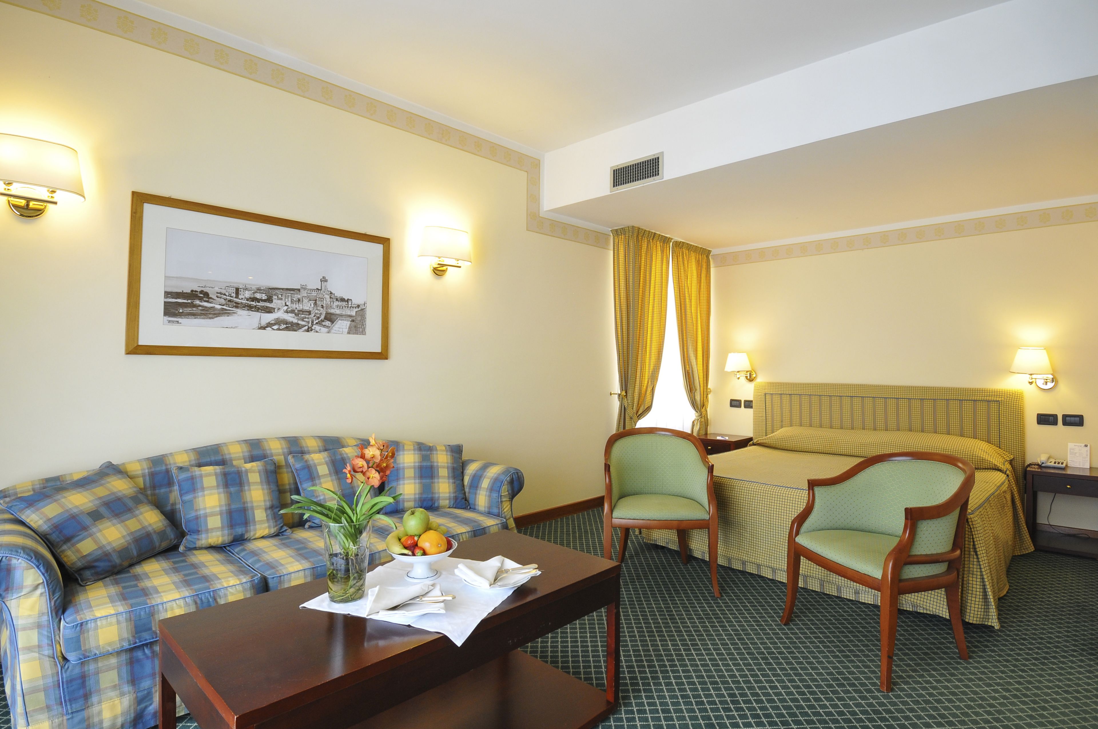 A hotel room with a checkered sofa, two armchairs, a wooden table with a vase of flowers, and a bed with a green bedspread.