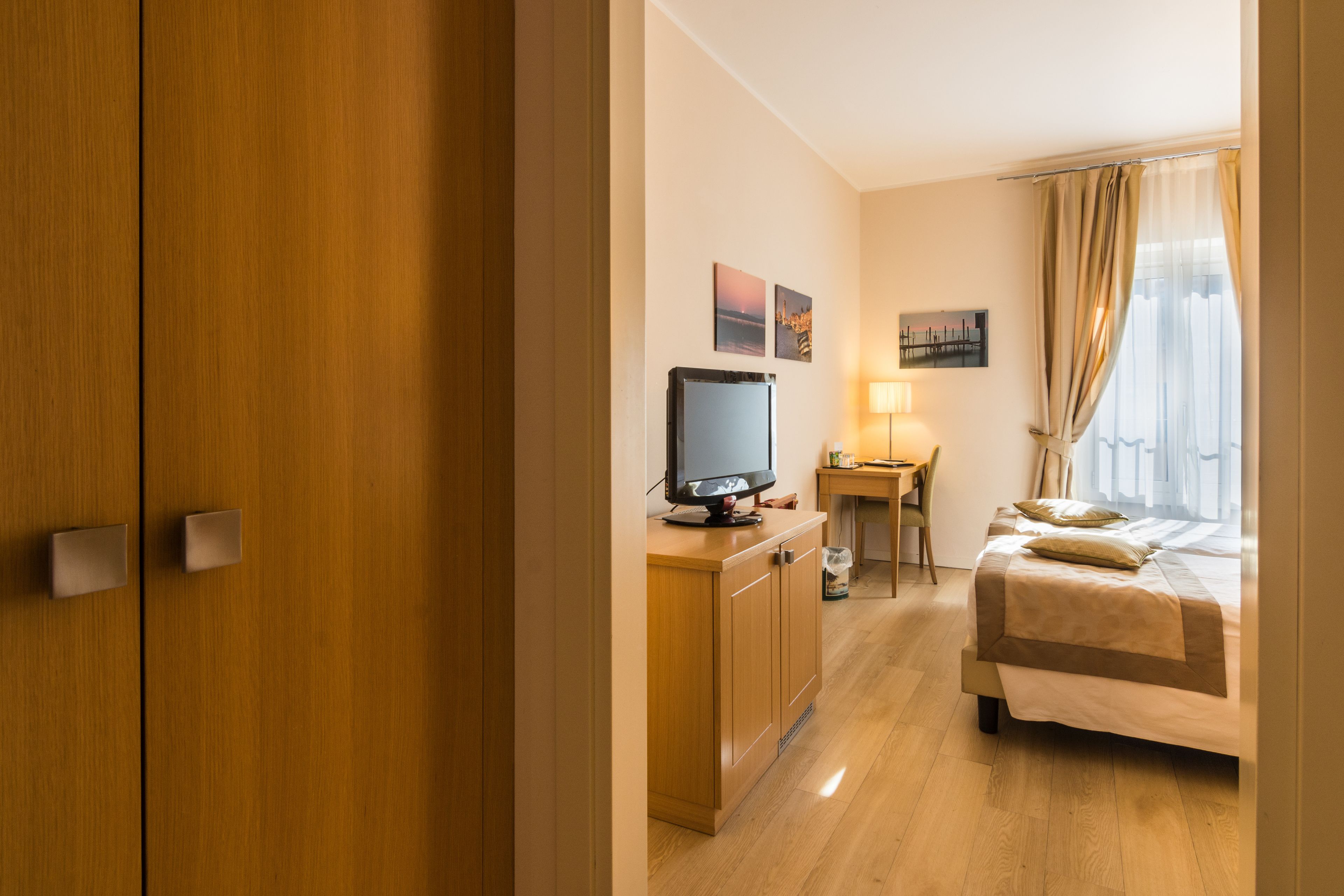 A hotel room with a wooden wardrobe, a desk with a computer, a bed with a beige bedspread, and a window with curtains.