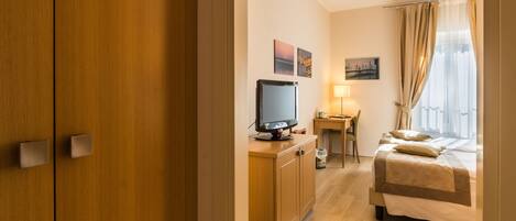 A hotel room with a wooden wardrobe, a desk with a computer, a bed with a beige bedspread, and a window with curtains.