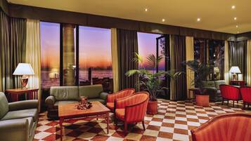 A hotel lobby with a checkered floor, leather sofas, armchairs, potted plants, and large windows offering a view of the sunset.