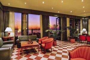 A hotel lobby with a checkered floor, leather sofas, armchairs, potted plants, and large windows offering a view of the sunset.