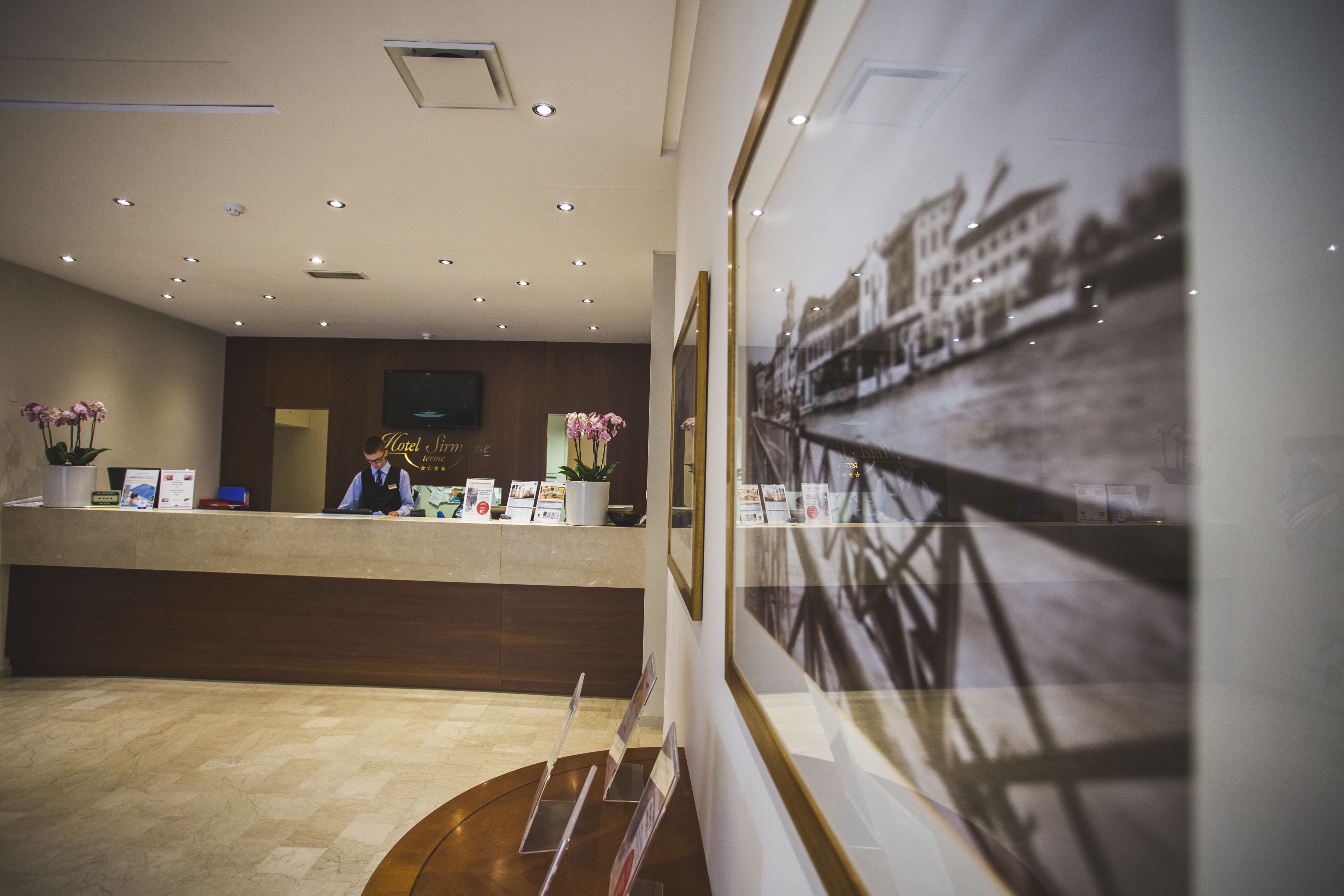 A hotel reception area with a wooden reception desk, a flat-screen TV, and framed photographs on the wall.