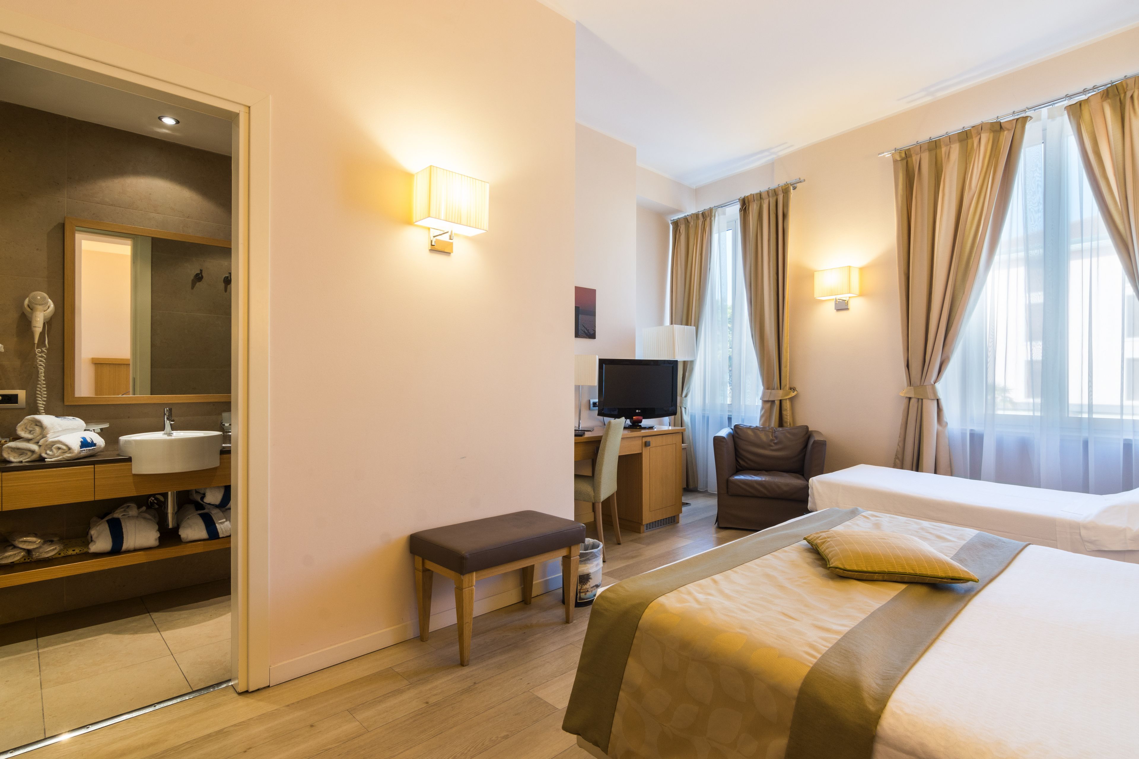 A hotel room with a bed, a desk, a chair, a TV, a bathroom with a sink, and a mirror.