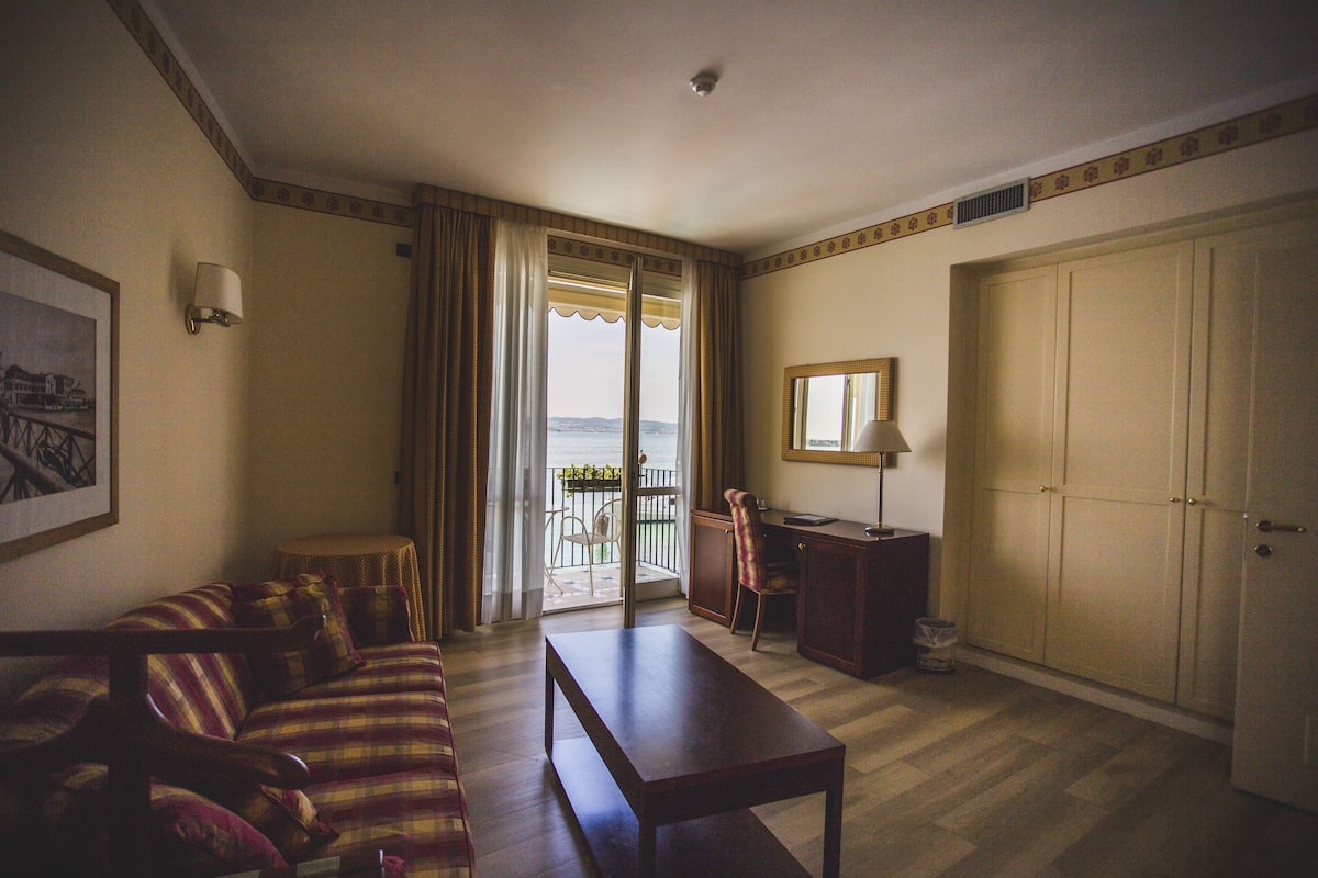 A living room with a sofa, a table, a chair, a lamp, a desk, and a wardrobe. There is a window with curtains and a view of the outdoors.