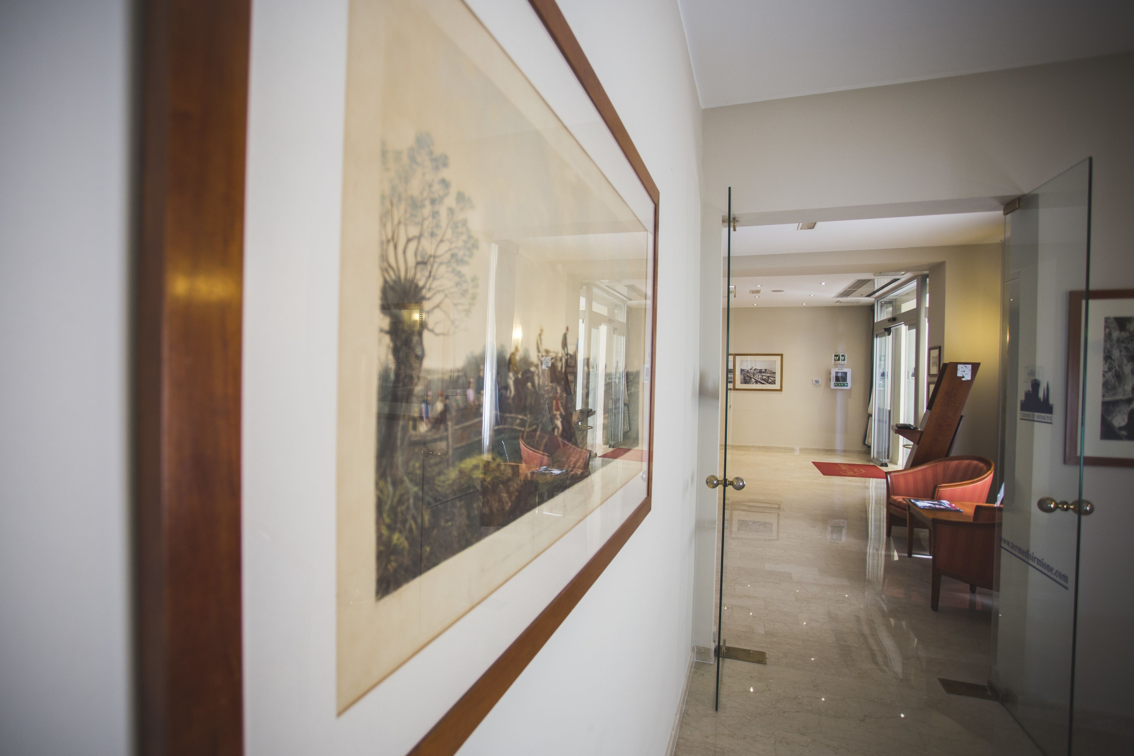 Un pasillo con una obra de arte grande enmarcadas en la pared, una puerta de vidrio que da a una habitación con una silla y una mesa, y un cuadro en la pared.