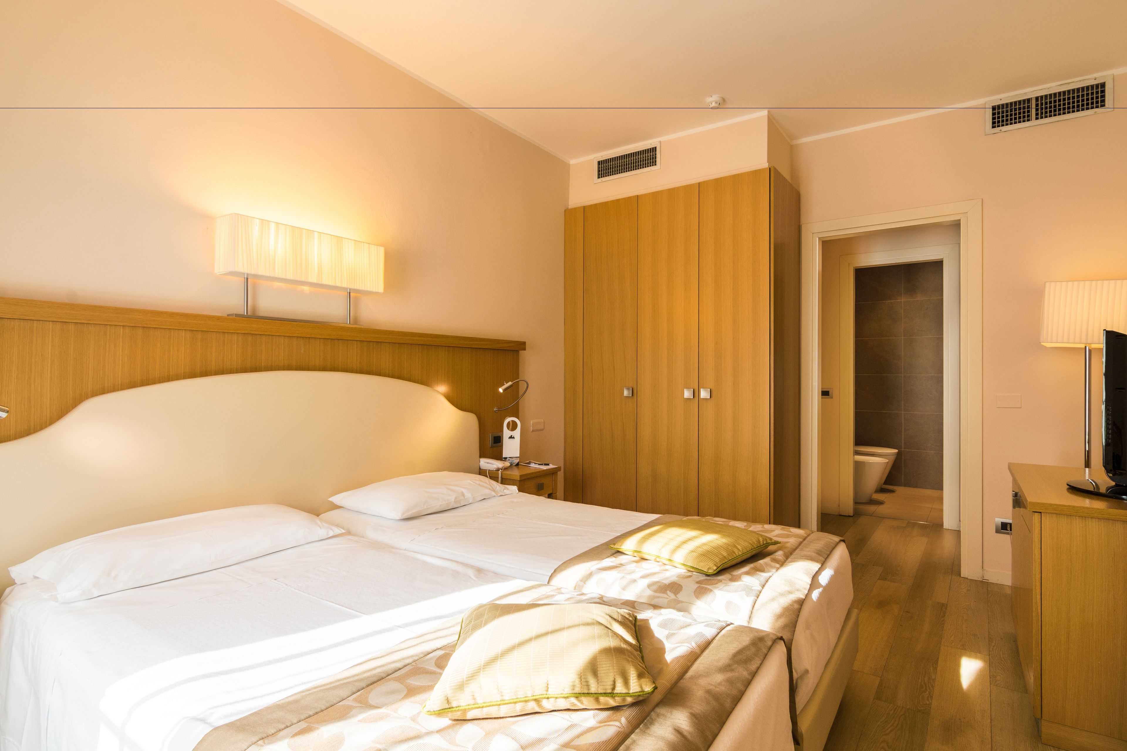 A hotel room with a large bed, a wooden wardrobe, a bedside table with a lamp, and a television.