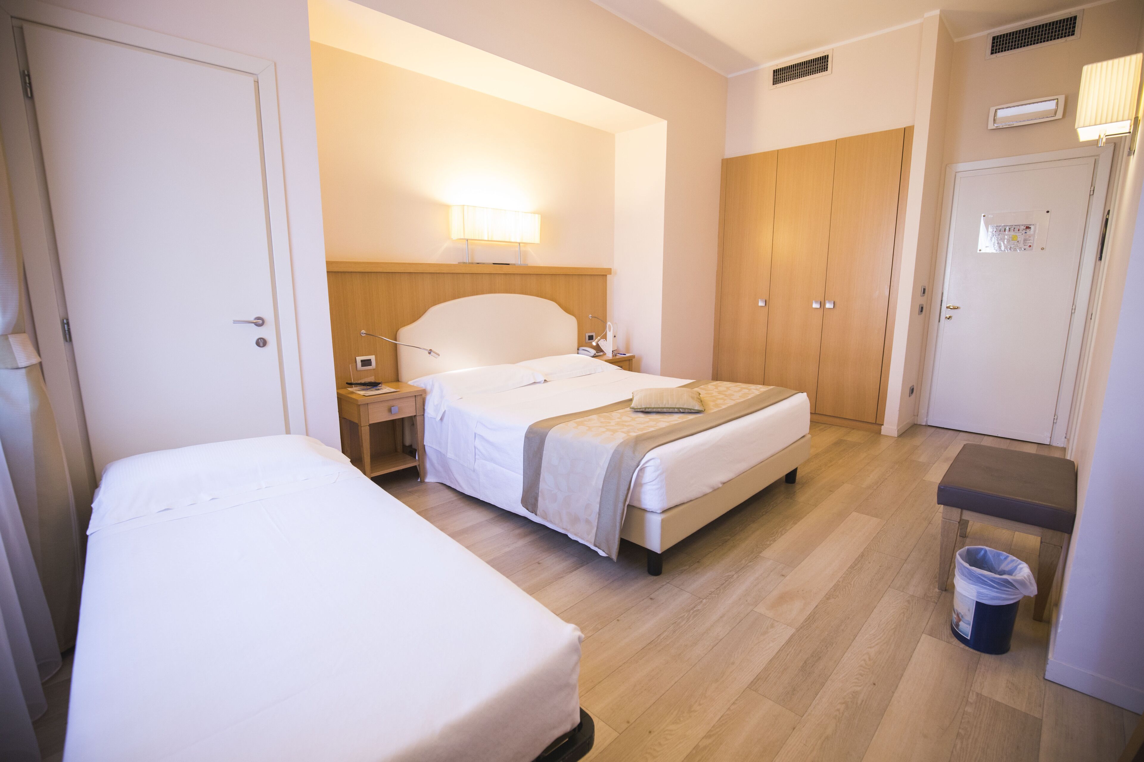 Une chambre d’hôtel avec deux lits, une table de chevet avec une lampe, une armoire et une poubelle.