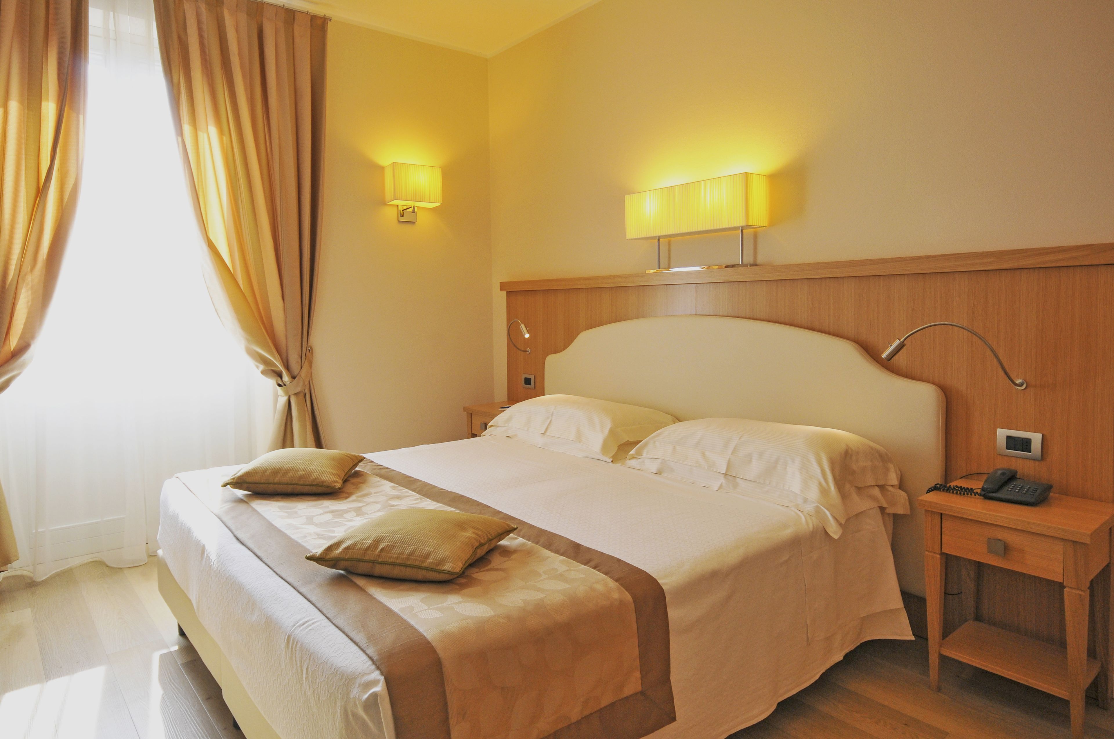 Habitación de hotel con una cama grande, dos lámparas de noche, una mesita con un teléfono y cortinas en la ventana.