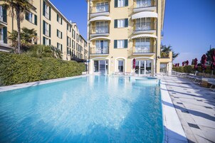 A swimming pool adjacent to a multi-story hotel with balconies, a well-maintained hedge, and sun loungers.