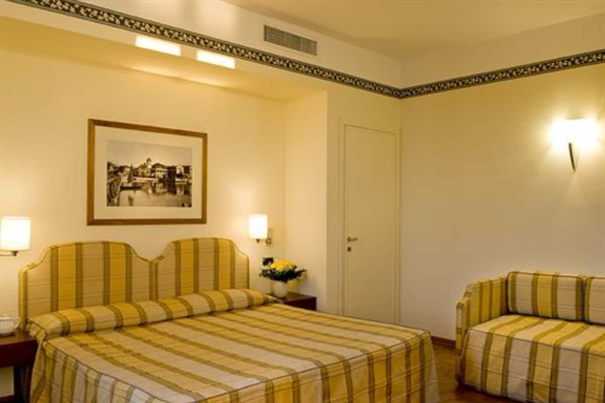 A hotel room with a bed, a bedside table, a lamp, a framed picture on the wall, and a door.
