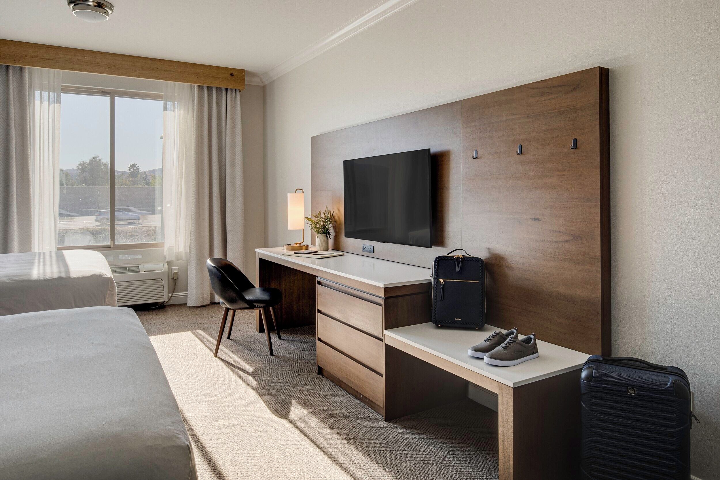 A hotel room with a bed, a desk with a chair, a TV, a suitcase, and a window with curtains.