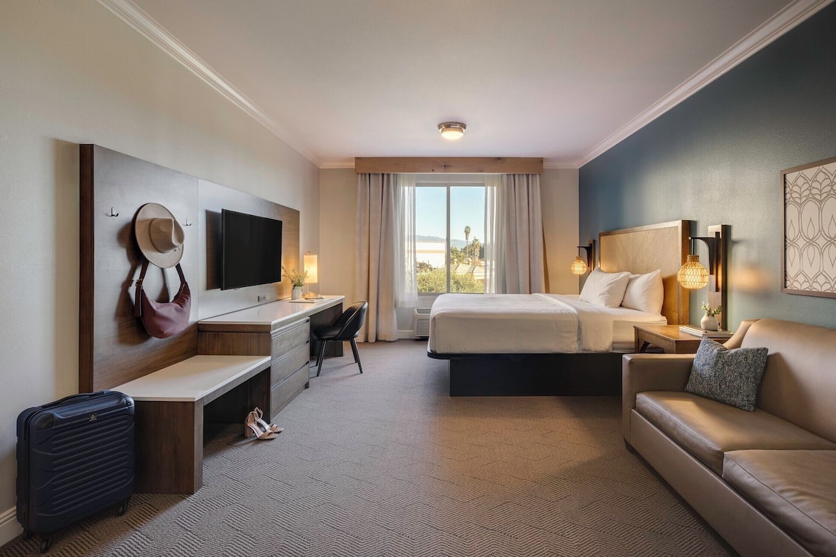 Ein Hotelzimmer mit Bett, Schreibtisch, Stuhl, Sofa, Fernseher und einem Fenster mit Vorhängen.