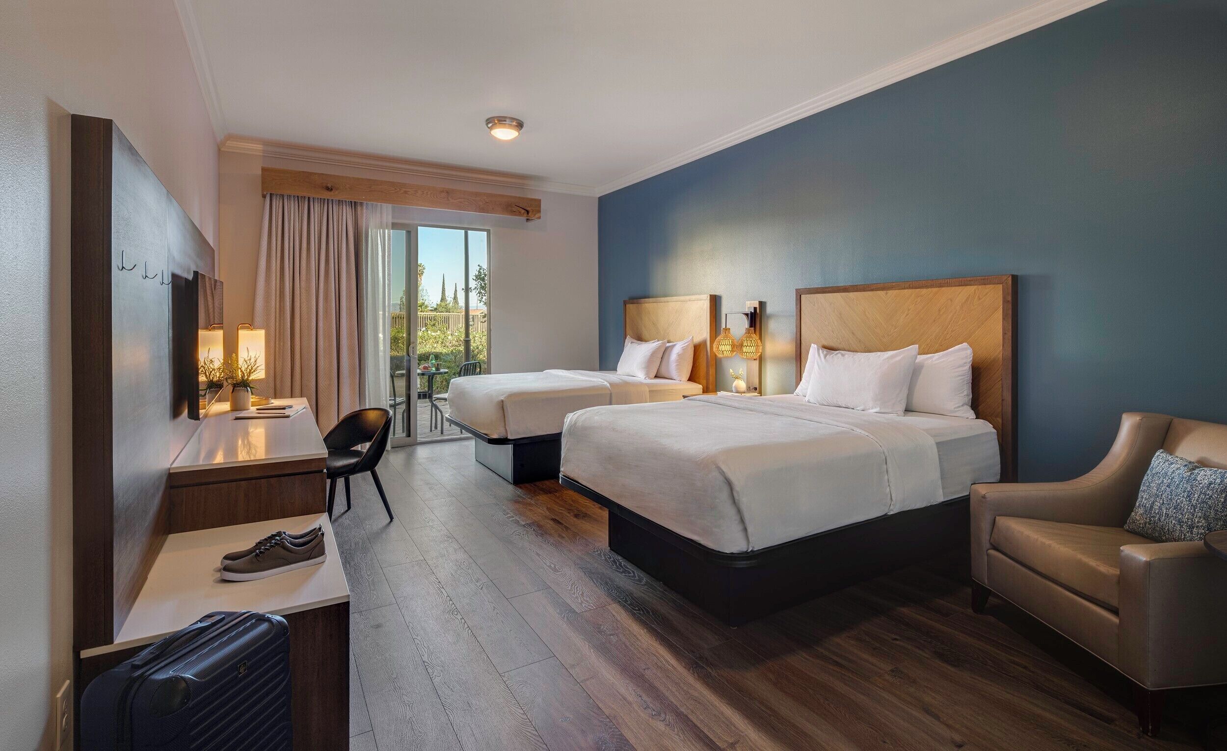 A hotel room with two beds, a desk, a chair, a sofa, and a view of the outdoors.