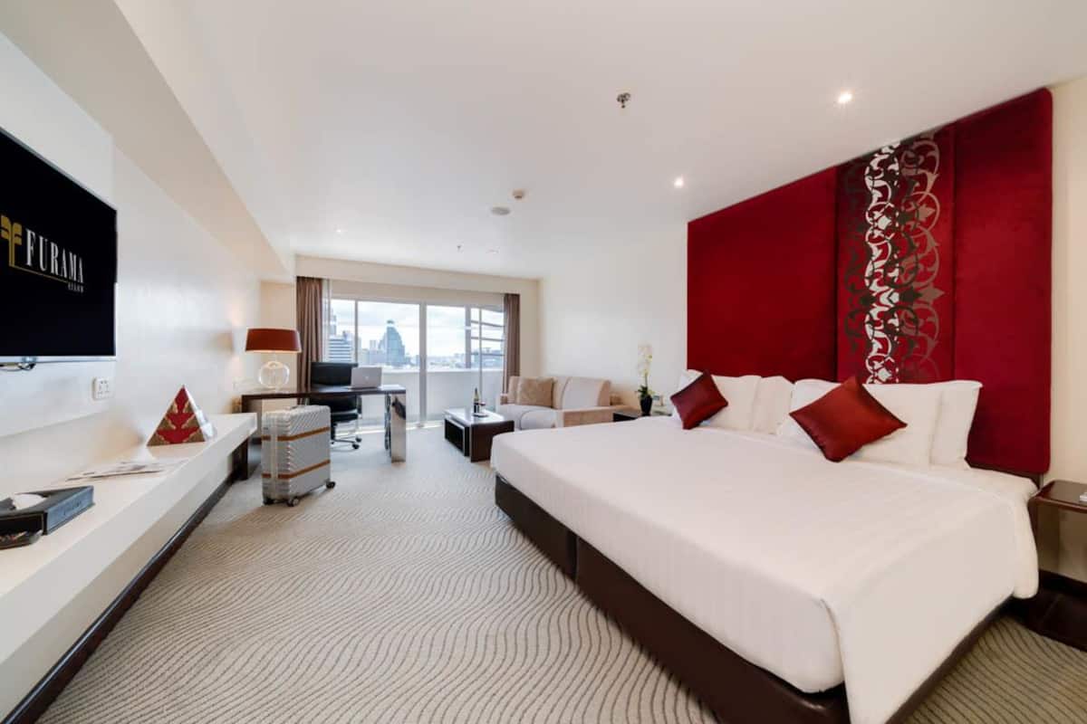 Ein Hotelzimmer mit einem großen Bett, einem Schreibtisch, einem Sessel, einem Fernseher und einem Fenster mit Blick auf die Stadt.