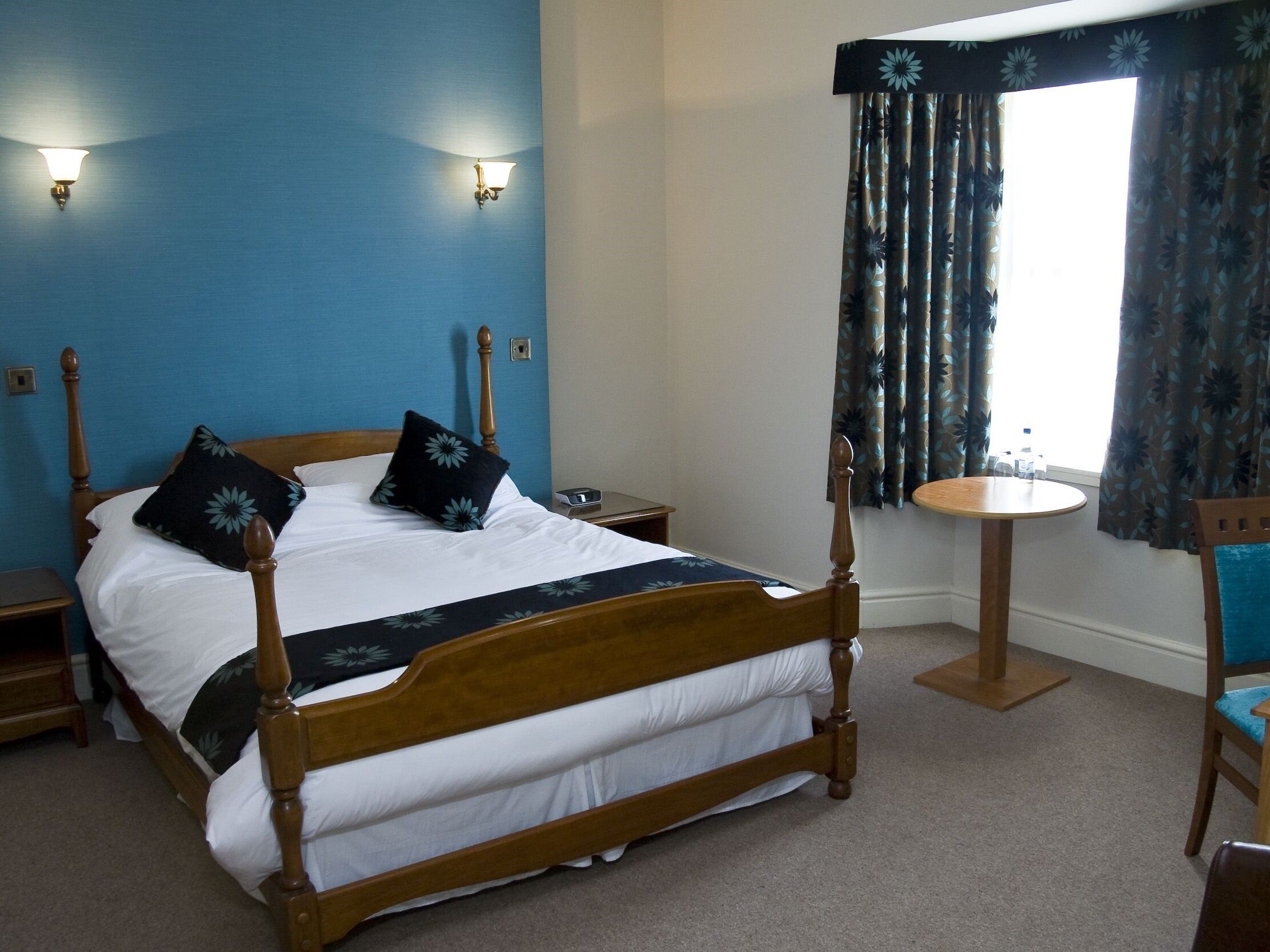 A neatly made bed with a wooden frame and white bedding, a bedside table with a lamp, a small round table with a chair, and a window with curtains.