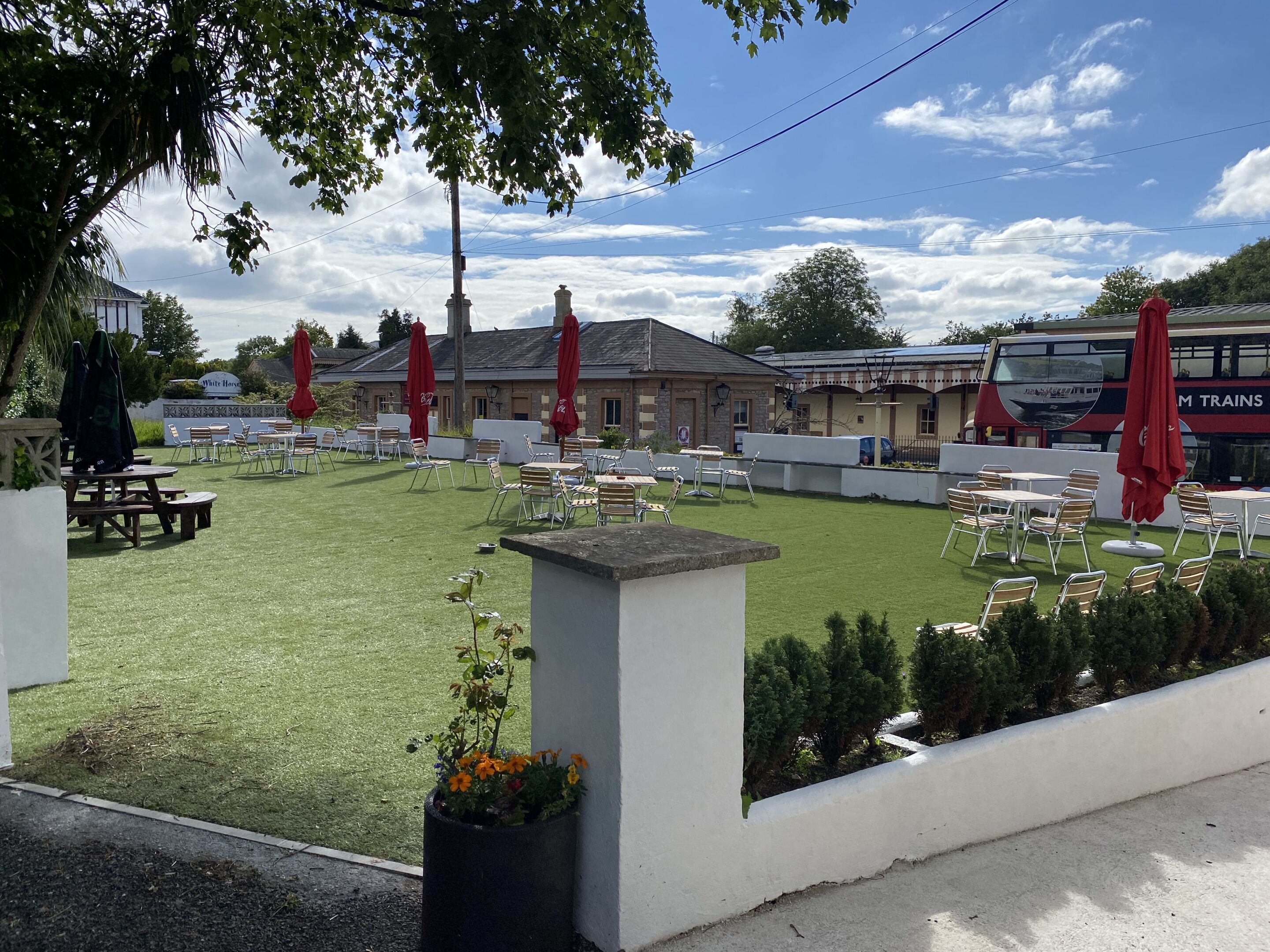 Un área de estar al aire libre con mesas y sillas, rodeada por un muro blanco y un edificio con un letrero que dice "White Horse".