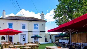 An outdoor seating area with wooden picnic tables and red umbrellas, set against a building with a white facade and a green lawn. - The Railway Inn (Brixham)
