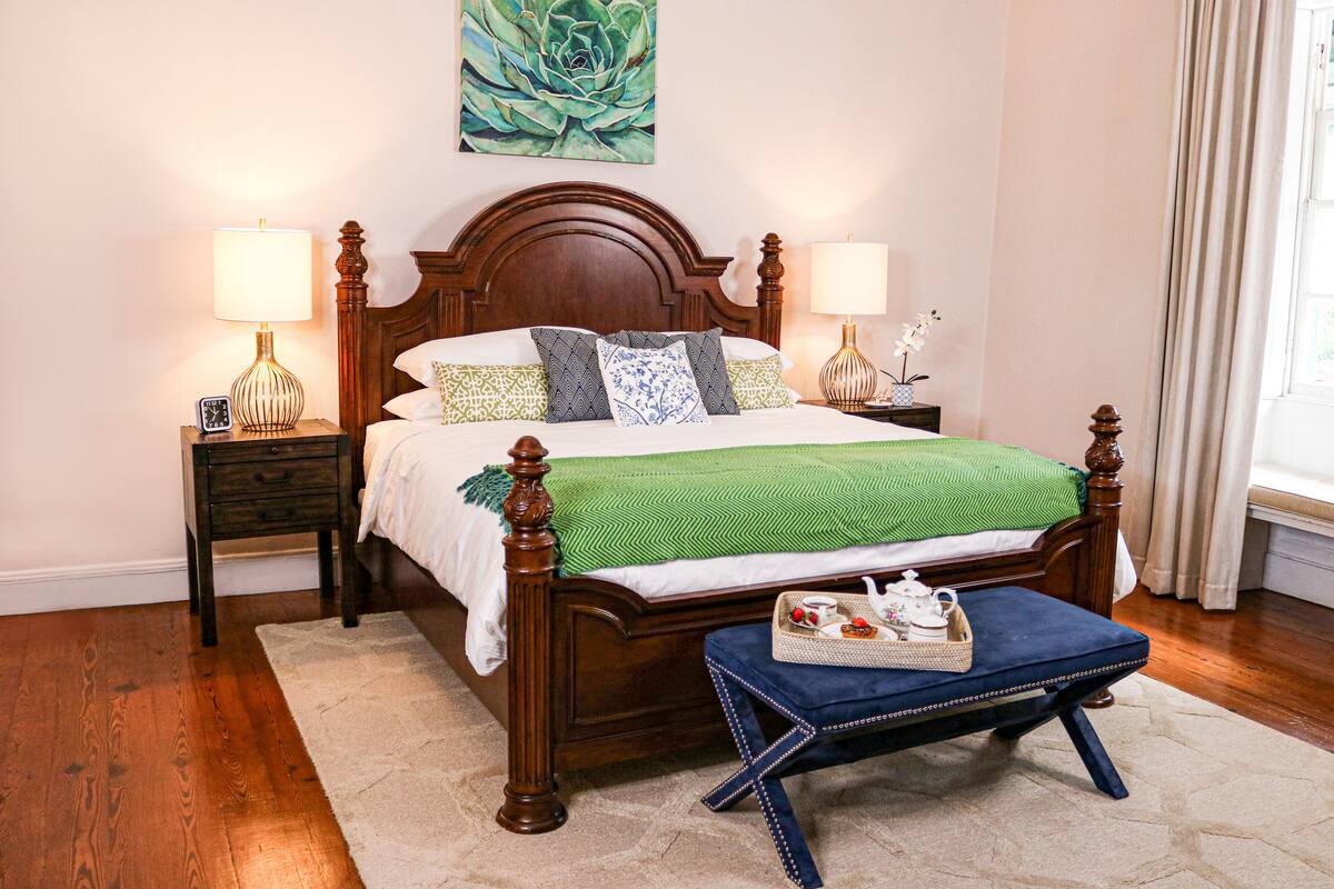 A neatly arranged bedroom with a wooden bed, two bedside tables with lamps, a decorative painting, and a window with curtains.