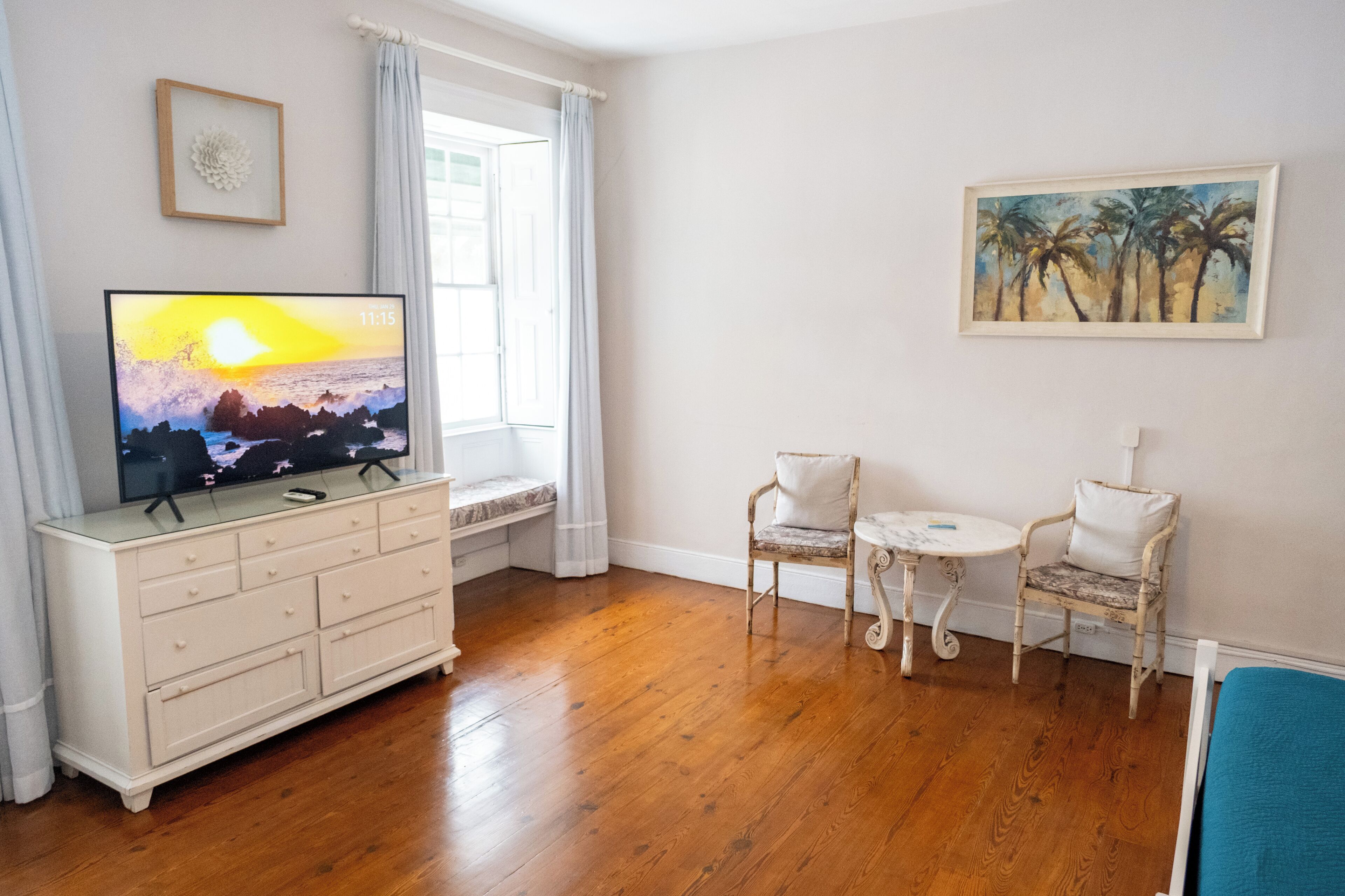 A living room with a wooden floor, a white entertainment unit with a TV, two chairs, a small table, and a painting on the wall.