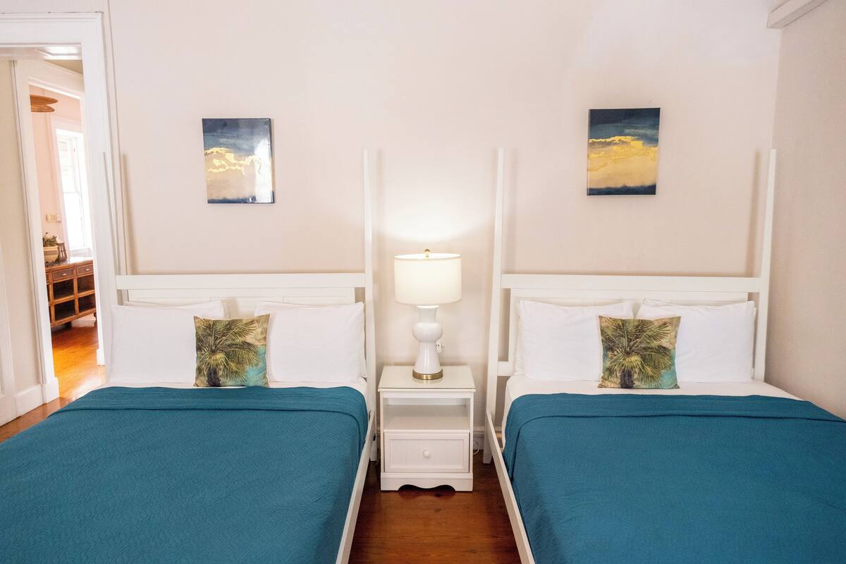 A hotel room with two beds, each with a teal bedspread and a palm tree pillow. There is a nightstand with a white lamp between the beds.