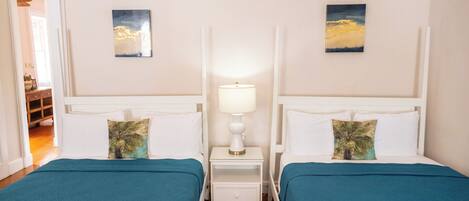 A hotel room with two beds, each with a teal bedspread and a palm tree pillow. There is a nightstand with a white lamp between the beds.