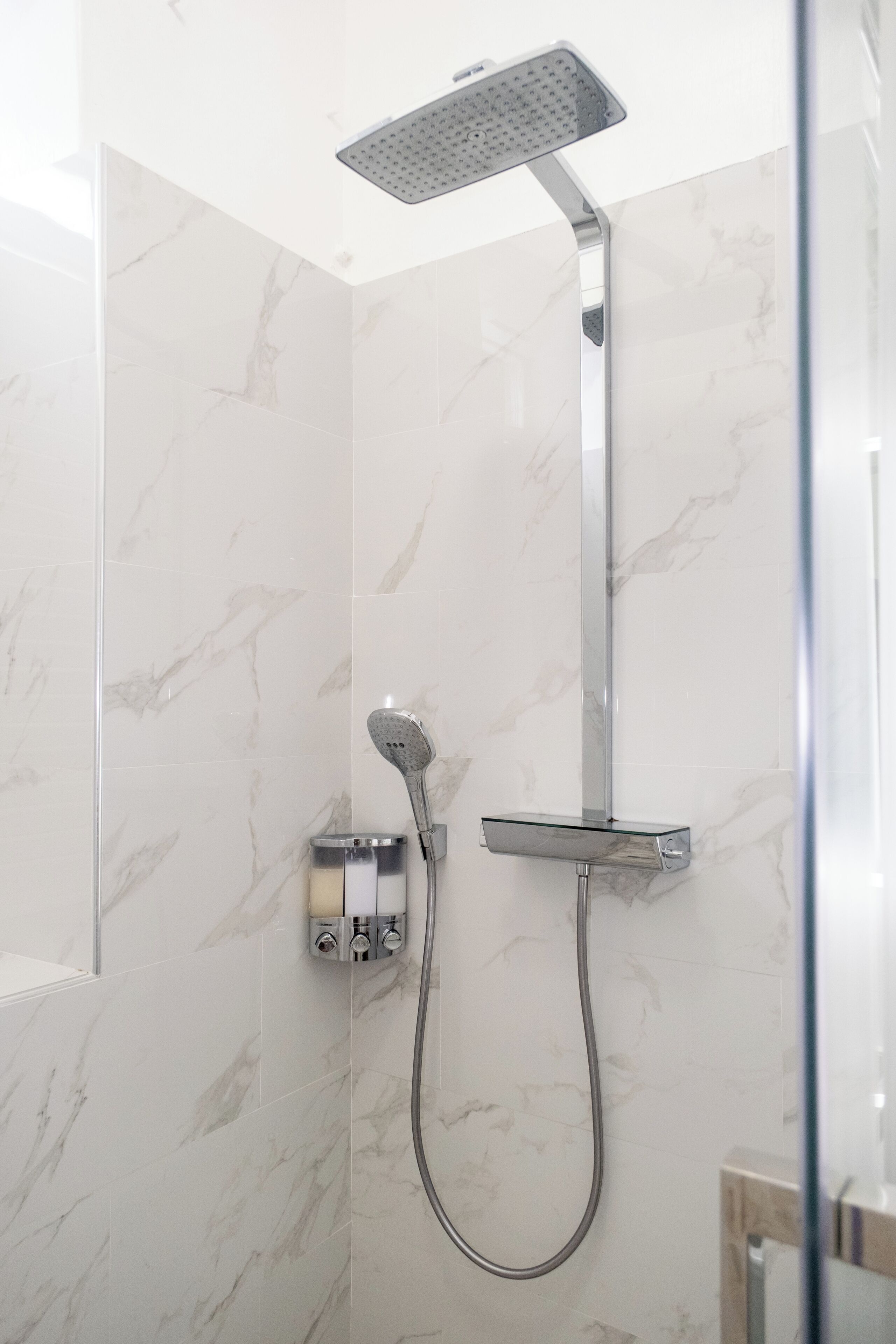 Ducha moderna con cabezal de ducha manual y otro fijo en el techo, instalada en una pared con azulejos de mármol.