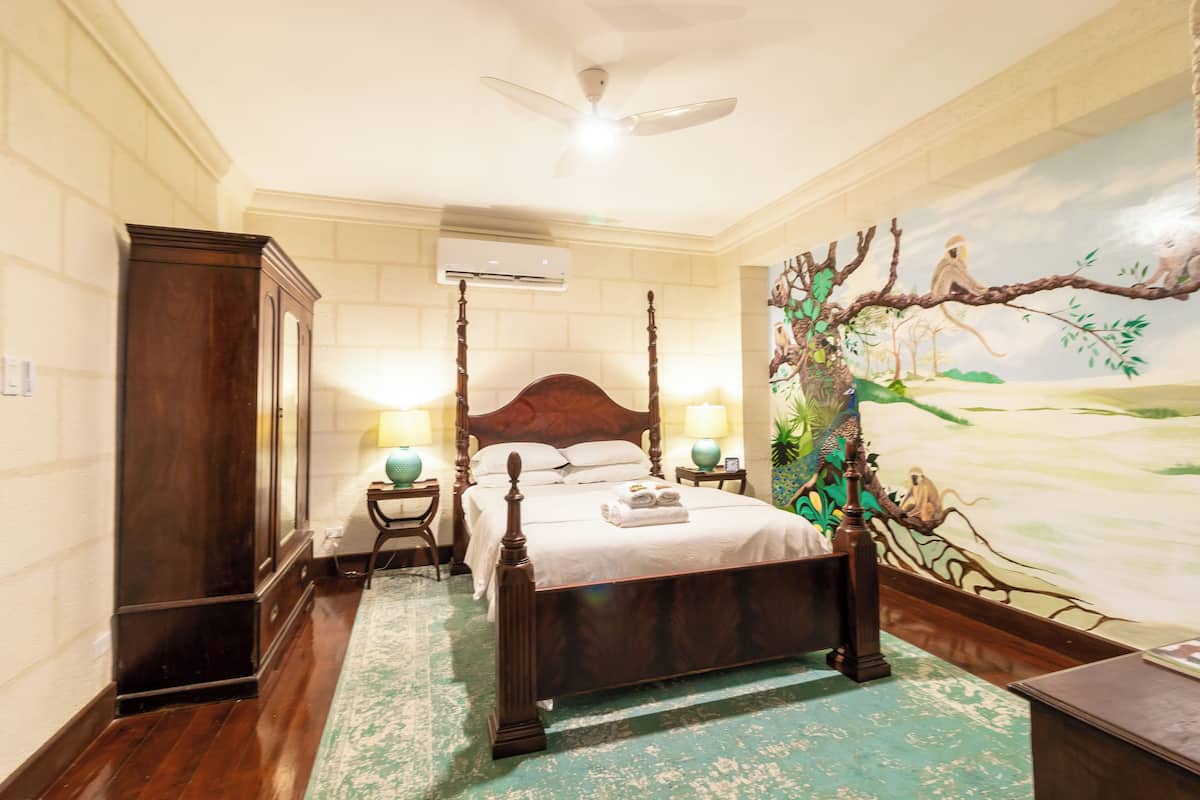 A bedroom with a four-poster bed, a ceiling fan, an air conditioning unit, a mural of a tree and animals, and a wooden wardrobe.