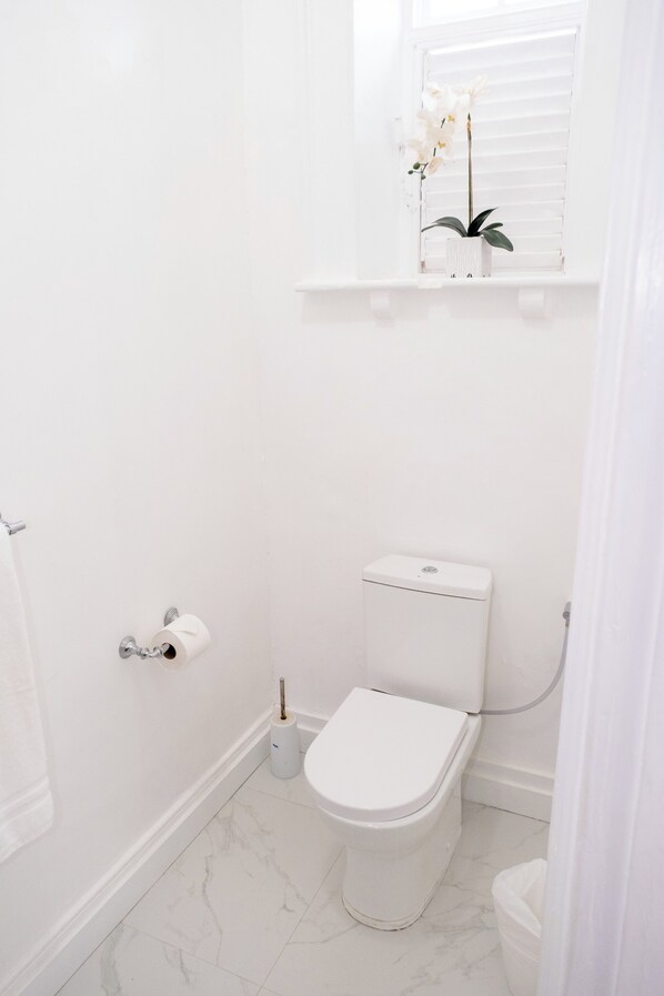 Un baño pequeño y blanco con inodoro, suelo de mármol, dispensador de papel higiénico y una ventana con una planta de orquídea.