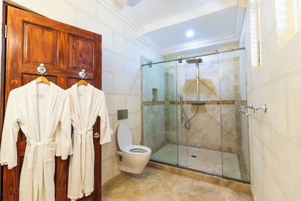 A bathroom with a wooden door, two white robes hanging on a rack, a toilet, and a glass-enclosed shower area. - Sweetfield Manor (Bridgetown)