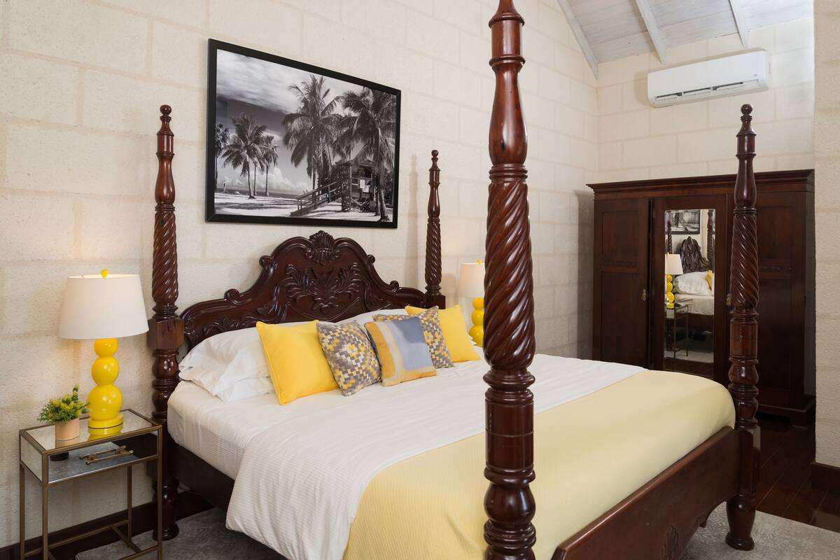 A four-poster bed with a white and yellow bedspread, a wooden headboard, and multiple pillows. There is a small bedside table with a lamp and a plant. A framed picture of palm trees is on the wall above the bed.
