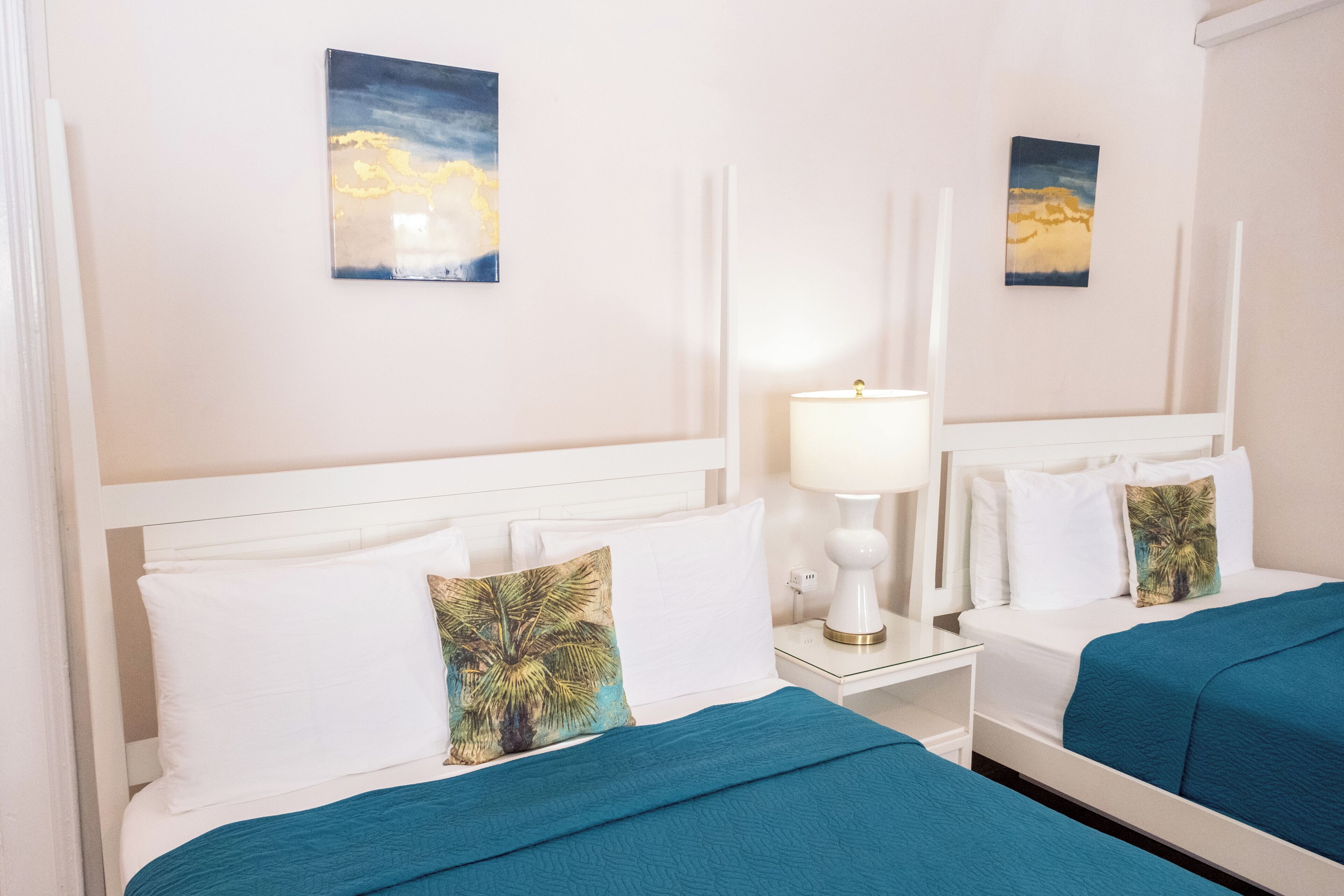 Una camera d'albergo con due letti, testiere bianche, biancheria da letto bianca, un comodino con una lampada e due quadri appesi al muro.