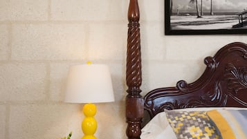 A neatly made bed with a patterned pillow, a bedside table with a yellow lamp, and a framed picture of palm trees on the wall.