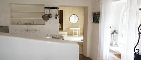 A kitchen with white walls, a wooden ceiling, and a window with white curtains. There are shelves with pots and pans, a round mirror, and a white radiator.