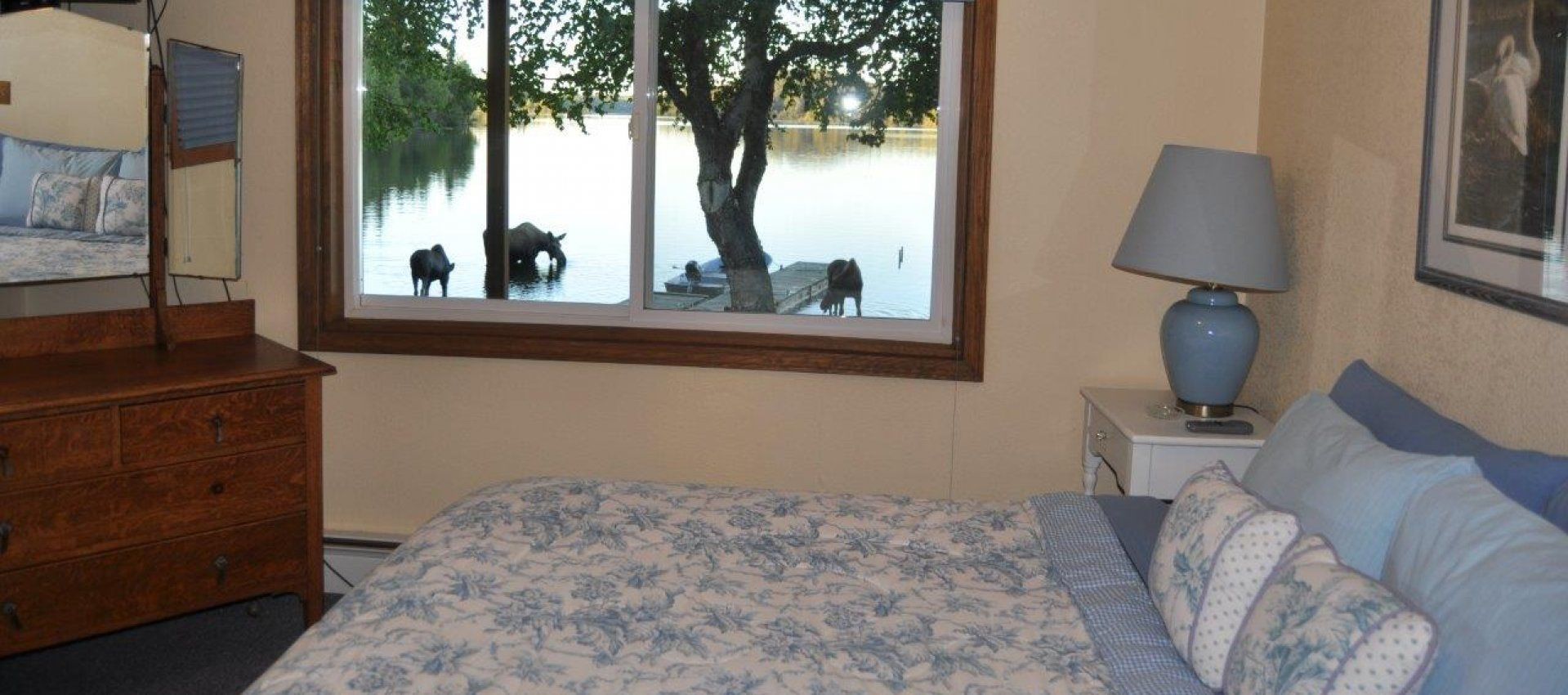 A bedroom with a bed, dresser, lamp, and a window with a view of a lake and moose.