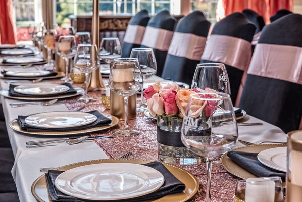 Een lange eettafel gedekt met borden, bestek en glazen, versierd met stoelen met roze en zwarte linten.