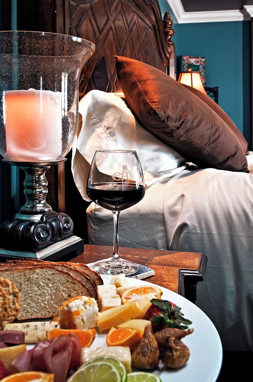 A cozy bedroom with a bed, pillows, a bedside table with a glass of red wine, a candle, and a plate of food including cheese, fruits, and bread.