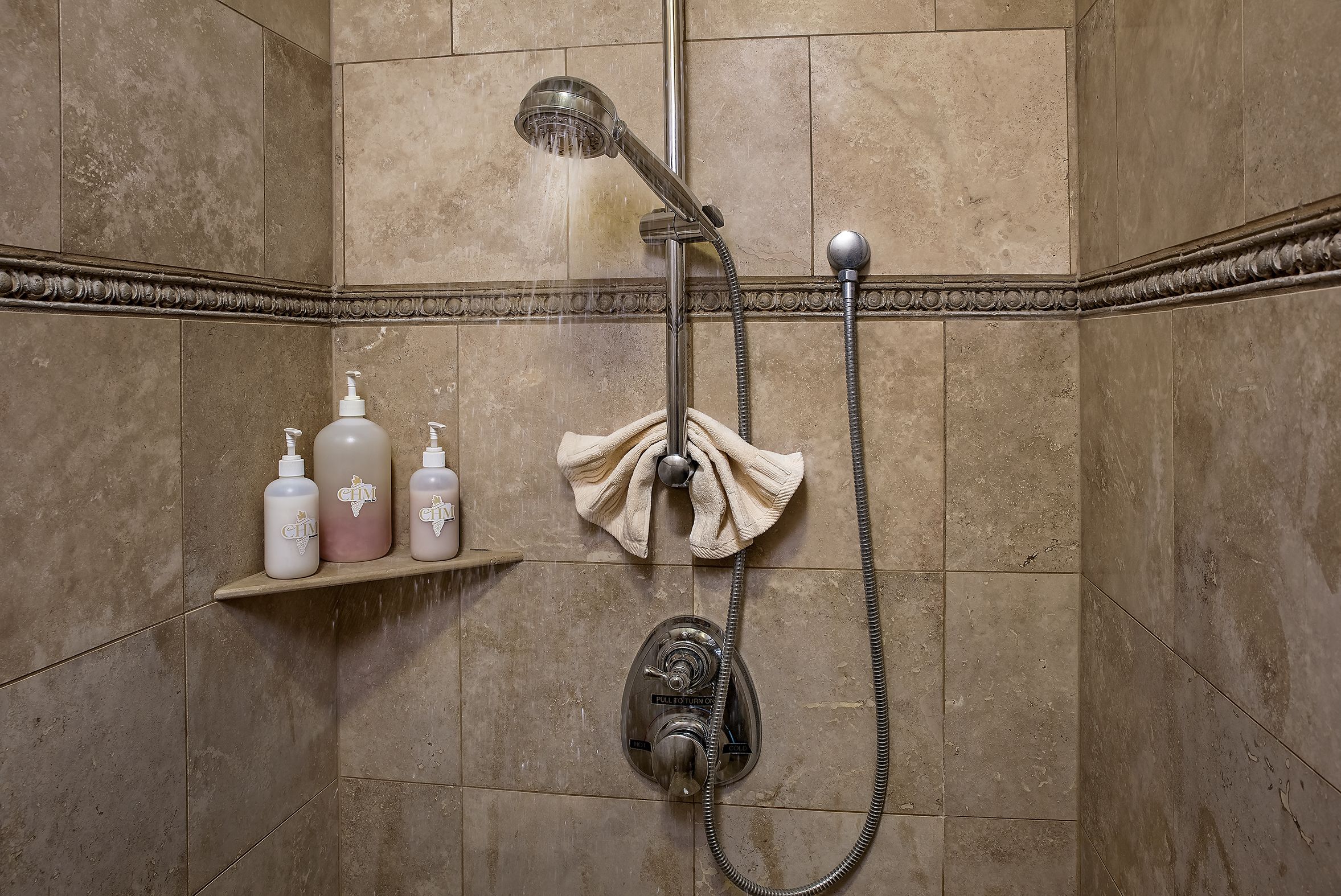 A shower with a handheld showerhead and a wall-mounted control valve. There are three bottles of shampoo on a shelf to the left.