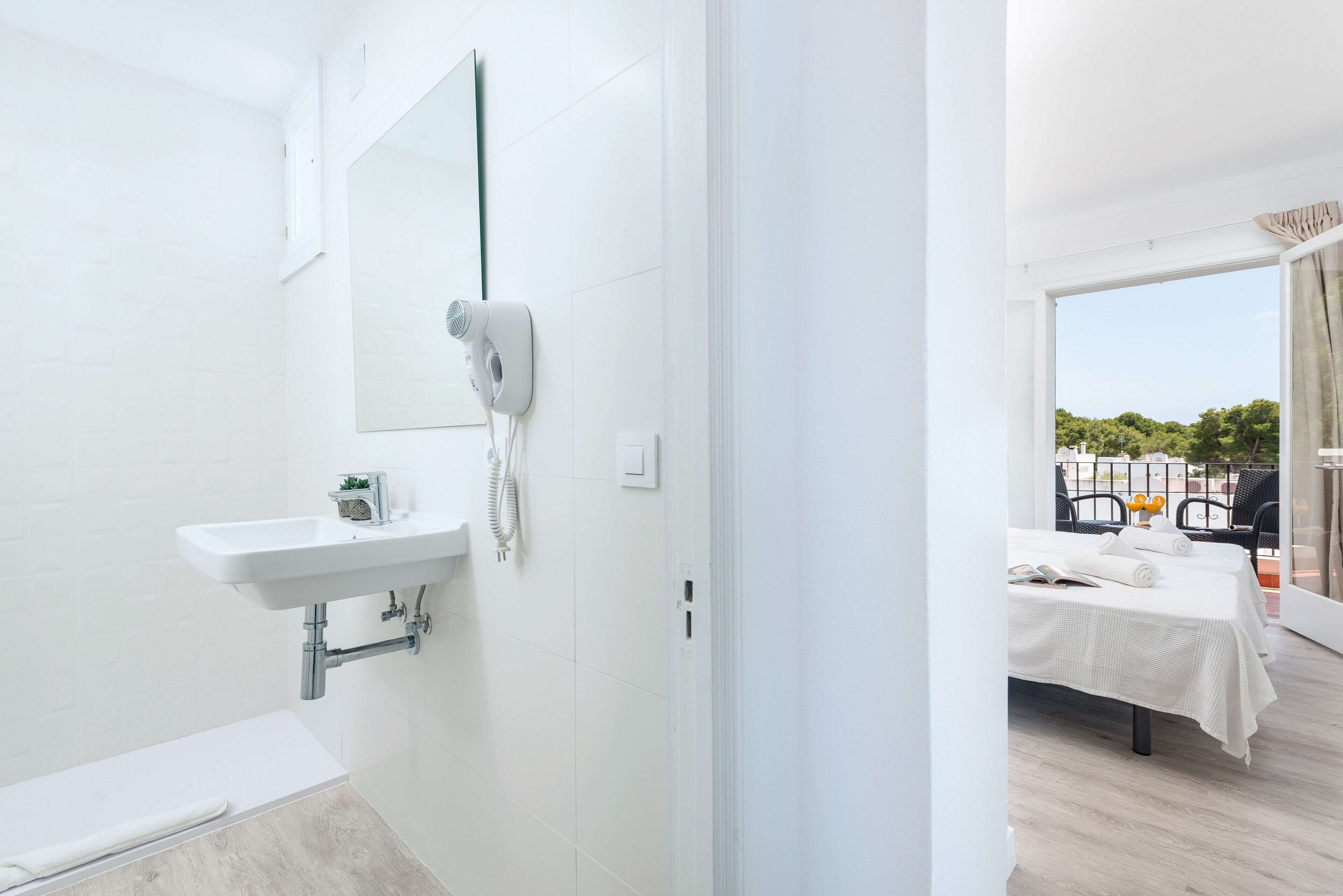 Casa de banho moderna com lavatório branco, espelho e secador de cabelo, com acesso a uma varanda com cama e cadeiras.