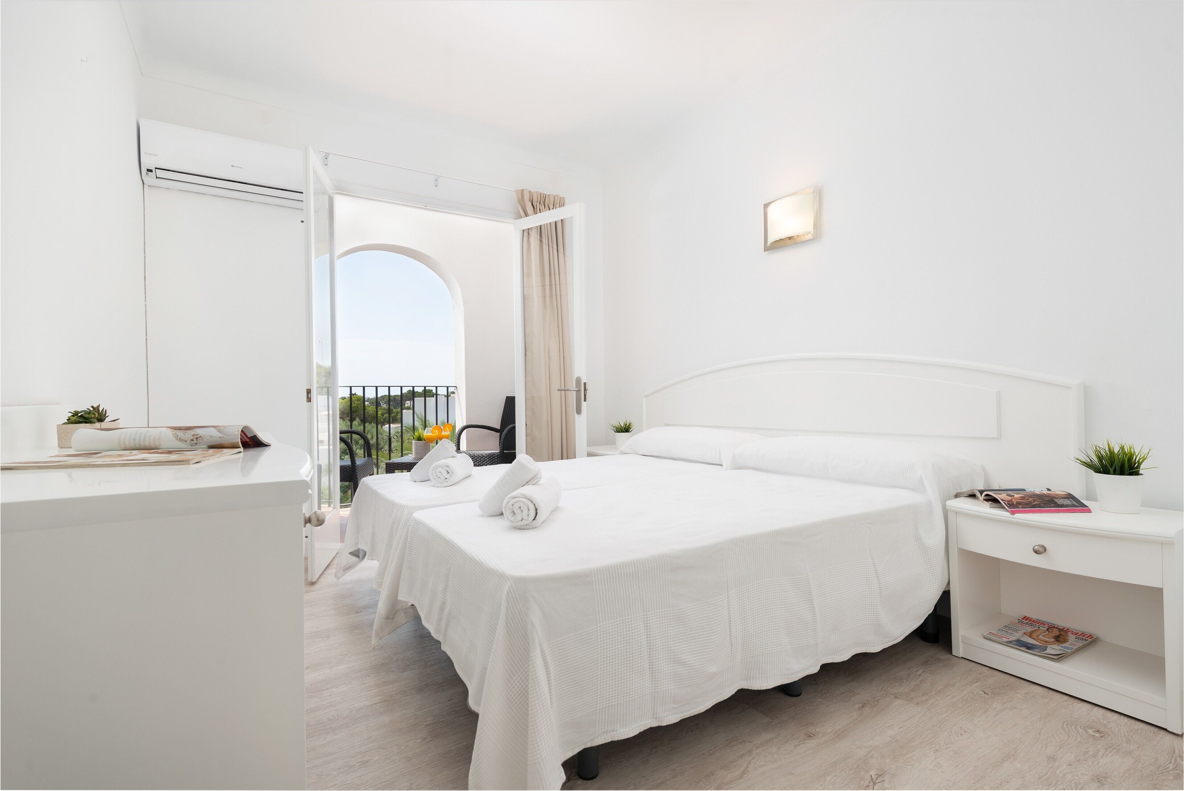 A hotel room with a large bed, a bedside table with a magazine, a small plant, and a view of the outdoors.