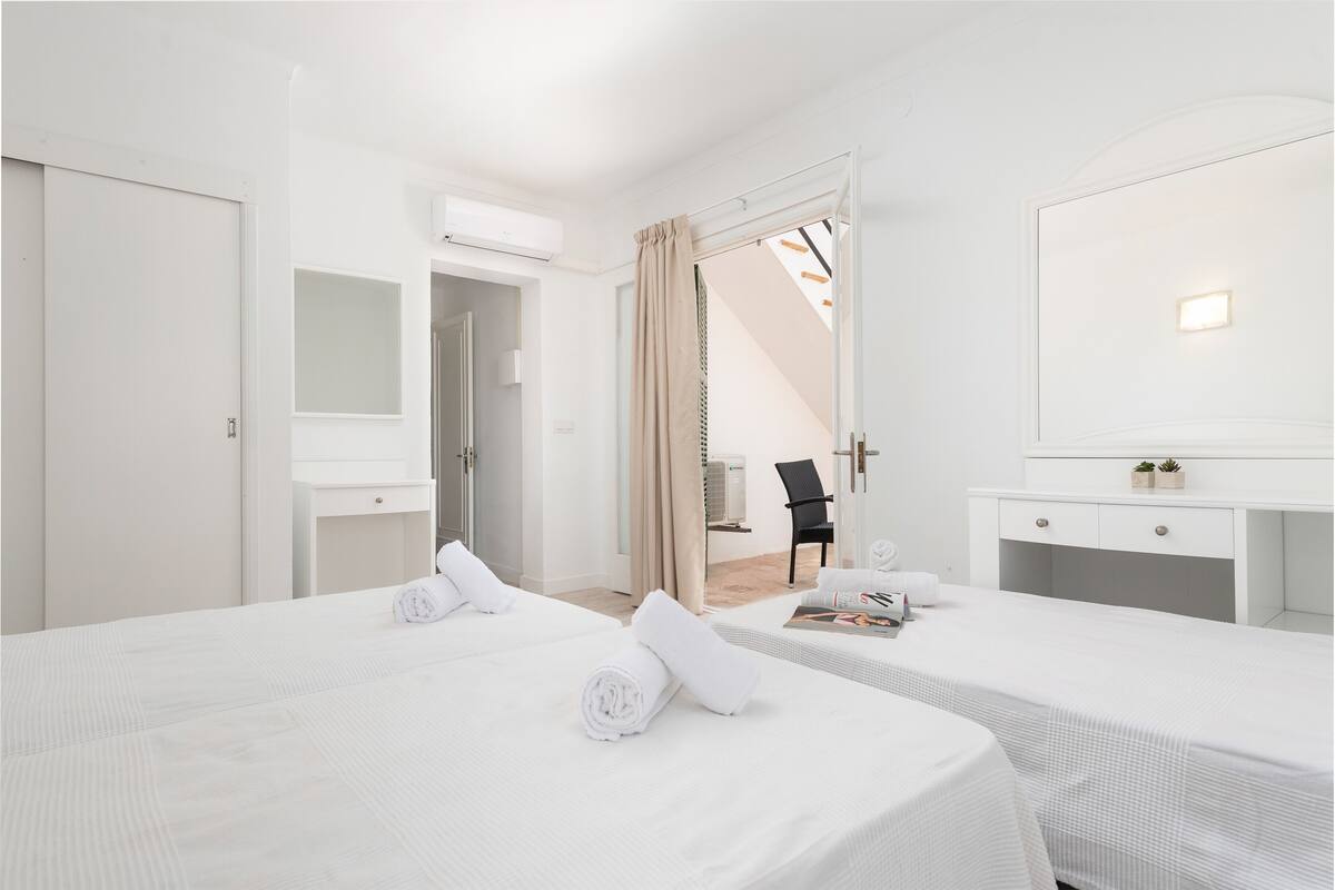 Ein Hotelzimmer mit zwei Betten, einem Schreibtisch und einem Sessel. Es gibt ein Fenster mit Vorhängen, eine Klimaanlage und eine kleine Pflanze auf dem Schreibtisch.
