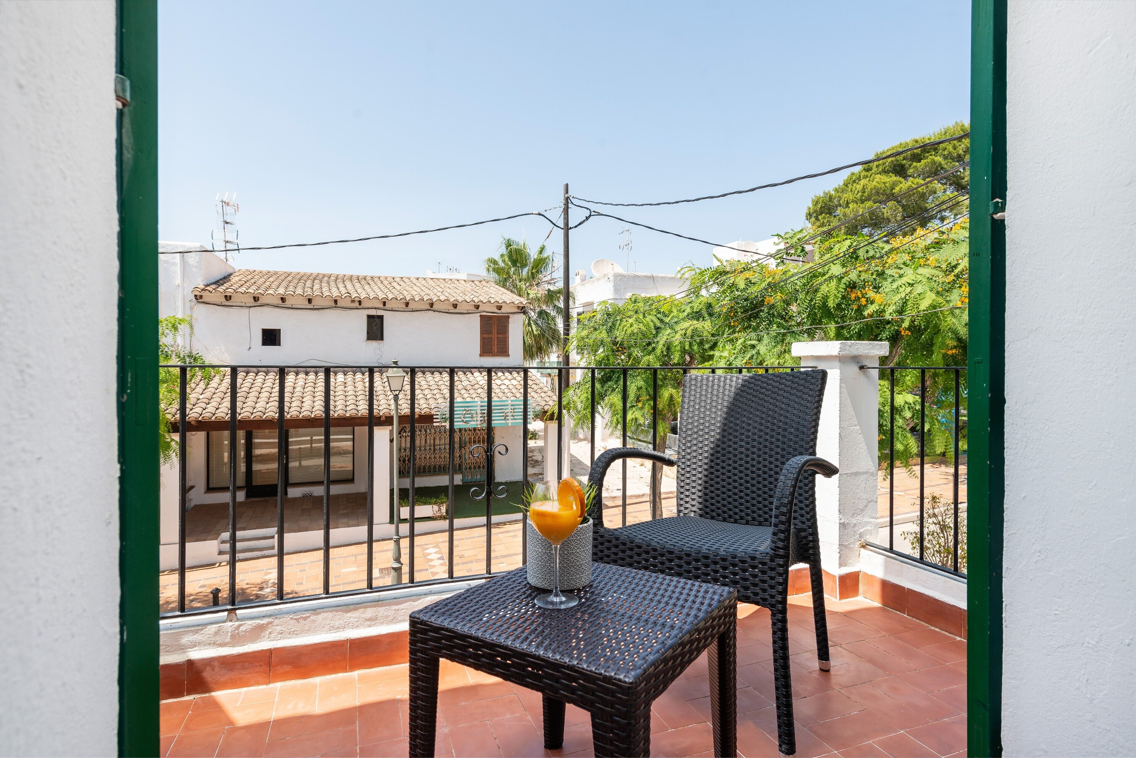 A balcony with a wicker chair and a table, a white wall, and a view of a building with a tiled roof.